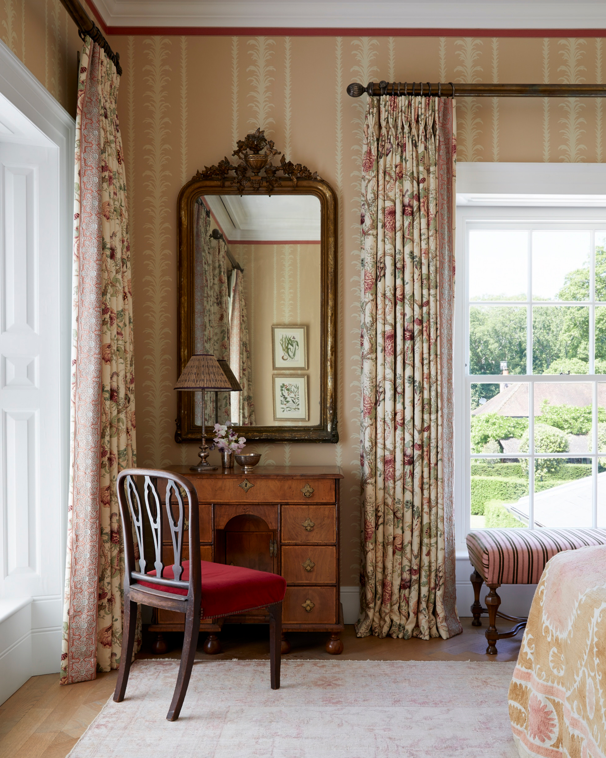a living room corner with an antique dressing table, an antique wooden chair with a red seat, an ornate gold mirror and a large window with floral curtains