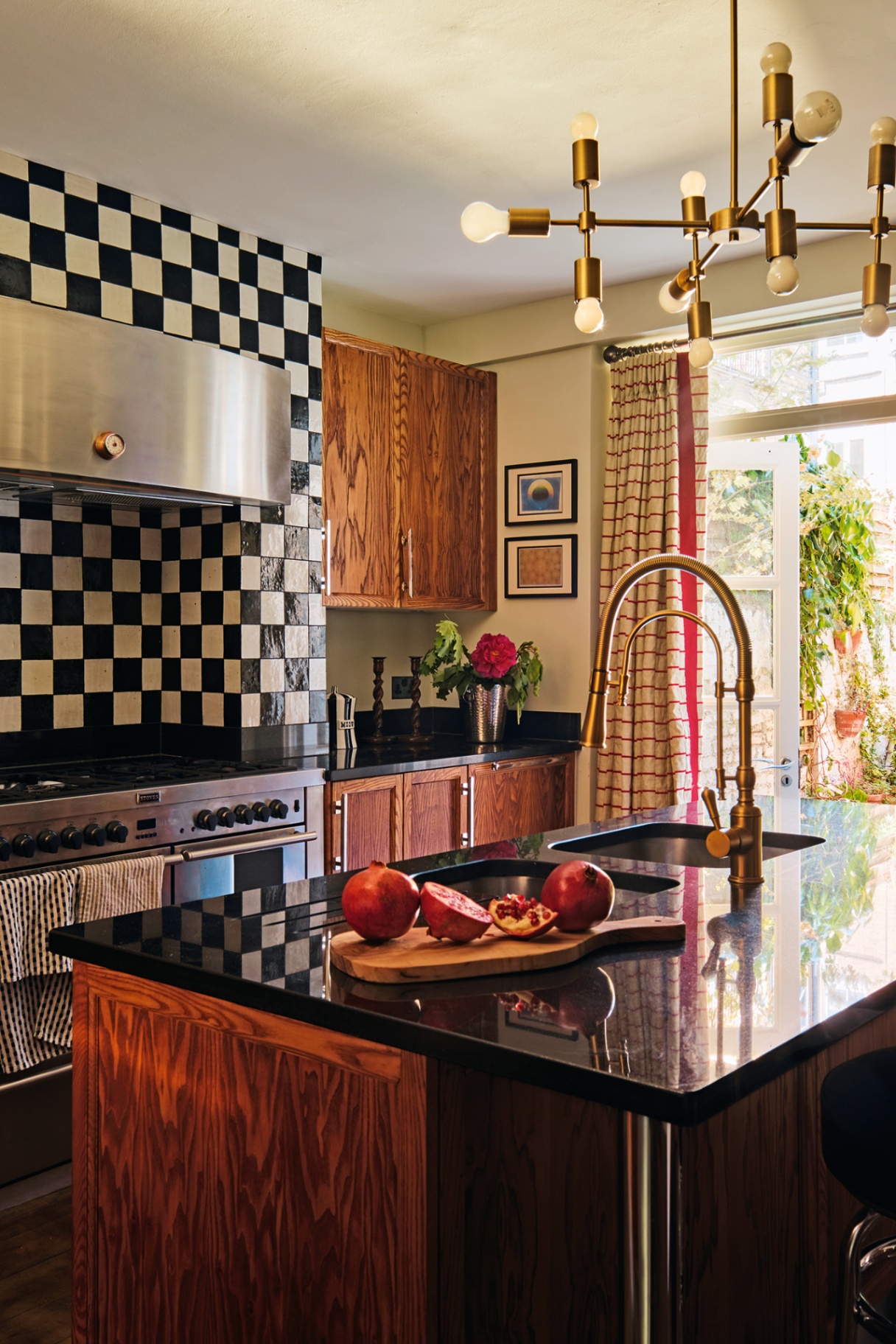 a kitchen with dark wood cabinets and black worktops. In the foreground is a large island with a sink, freestanding tap and pomegranates and in the back is a stove with a black-and-white tiled backsplash that reaches the ceiling