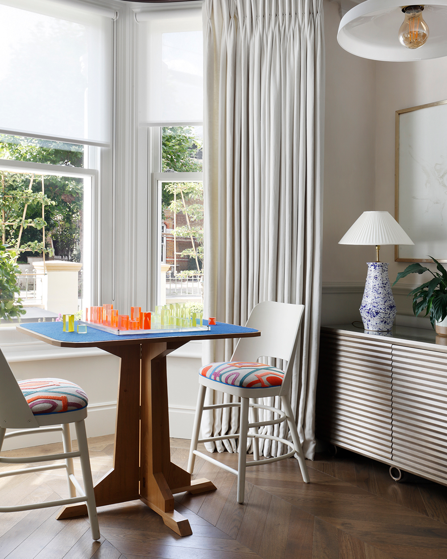 A blue and wood games table by a large window with white curtains, a ribbed side board with a blue and white lamp and a white pendant light above