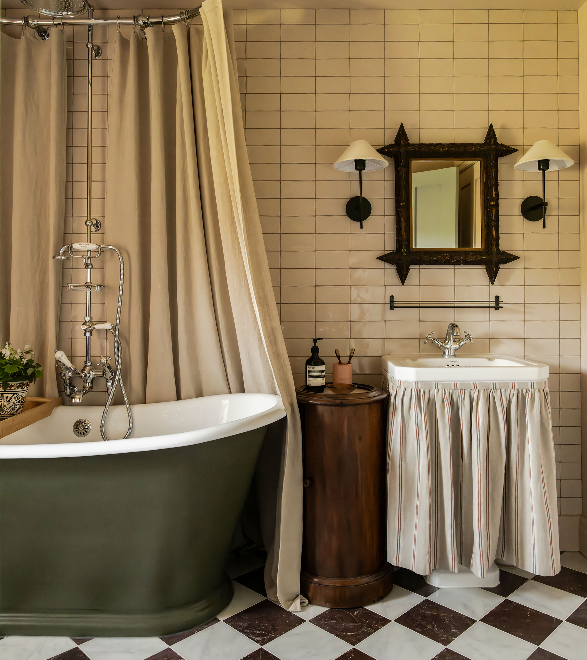 a bathroom with a dark green roll top bath with a silver shower and a canopy style shower curtain, a wooden unit next to it and a sink with a fabric skirt, a rectangular wooden mirror above and two sconces either side