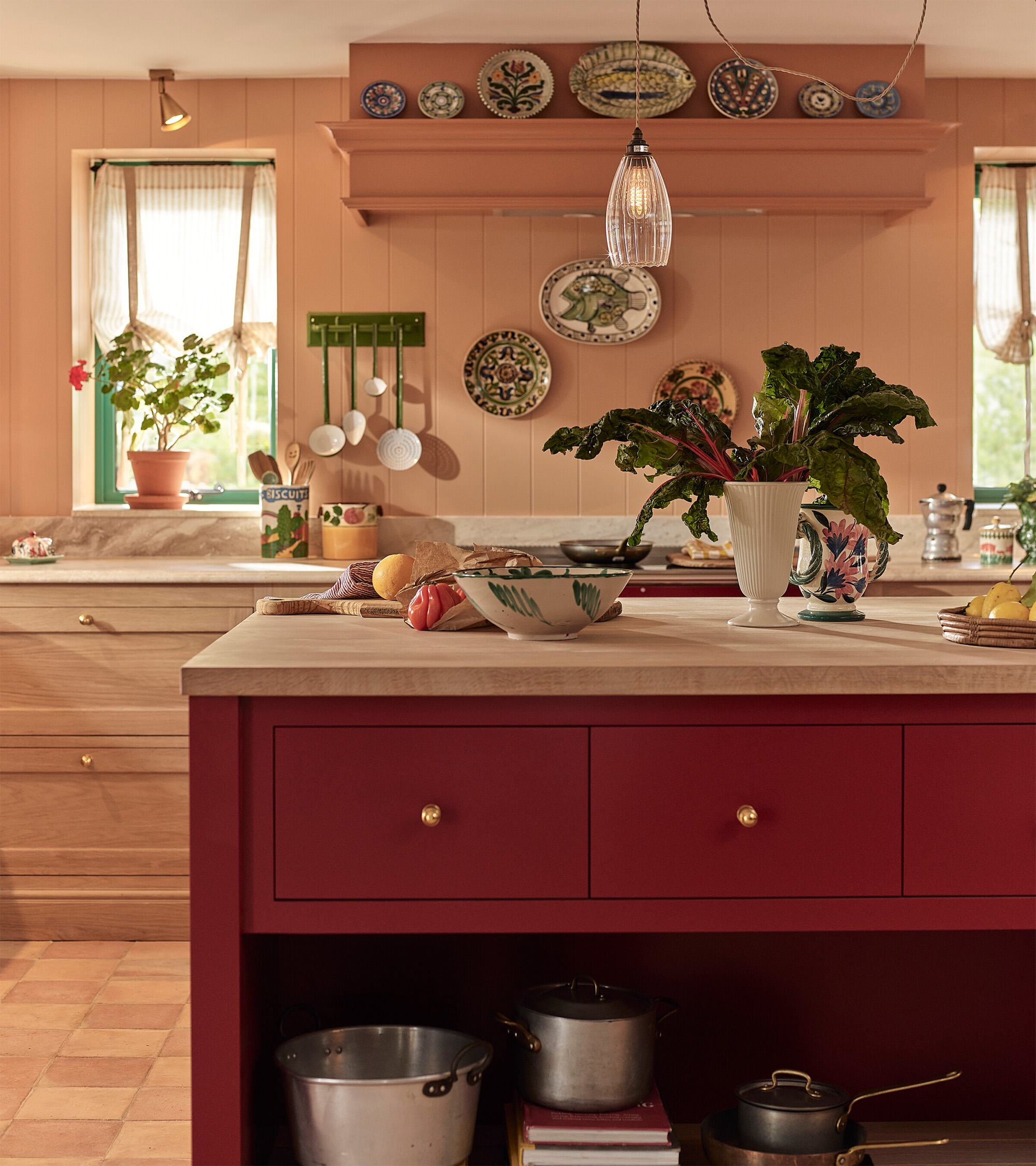 a kitchen with peach walls and decorative plates hung on the walls, a red kitchen island with a vase of rhubarb stalks and a clear glass pendant hanging above