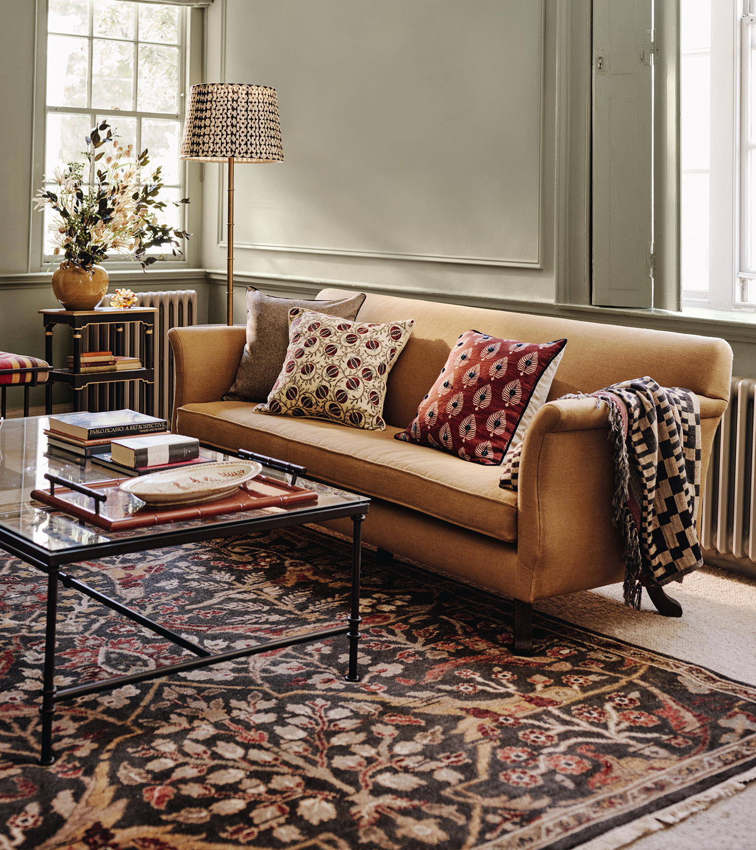A LIVING ROOM CORNER WITH sage green panelled walls, a mustard yellow sofa with patterned cushions, a rectangle metal coffee table and a large red patterened persian rug
