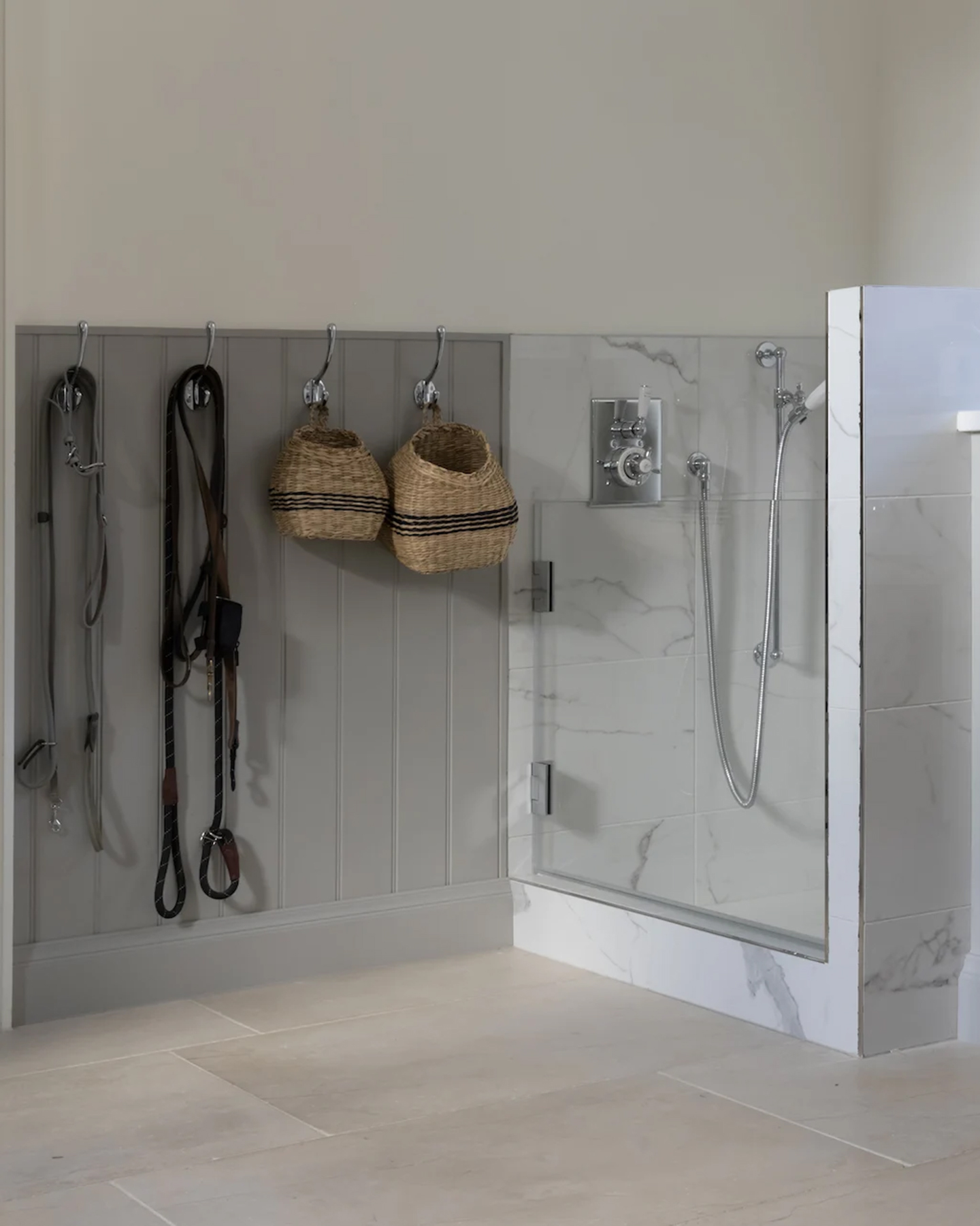 A utility room with a white marble dog shower zone and silver pegs on the wall with wicker baskets and dog leads hanging from them
