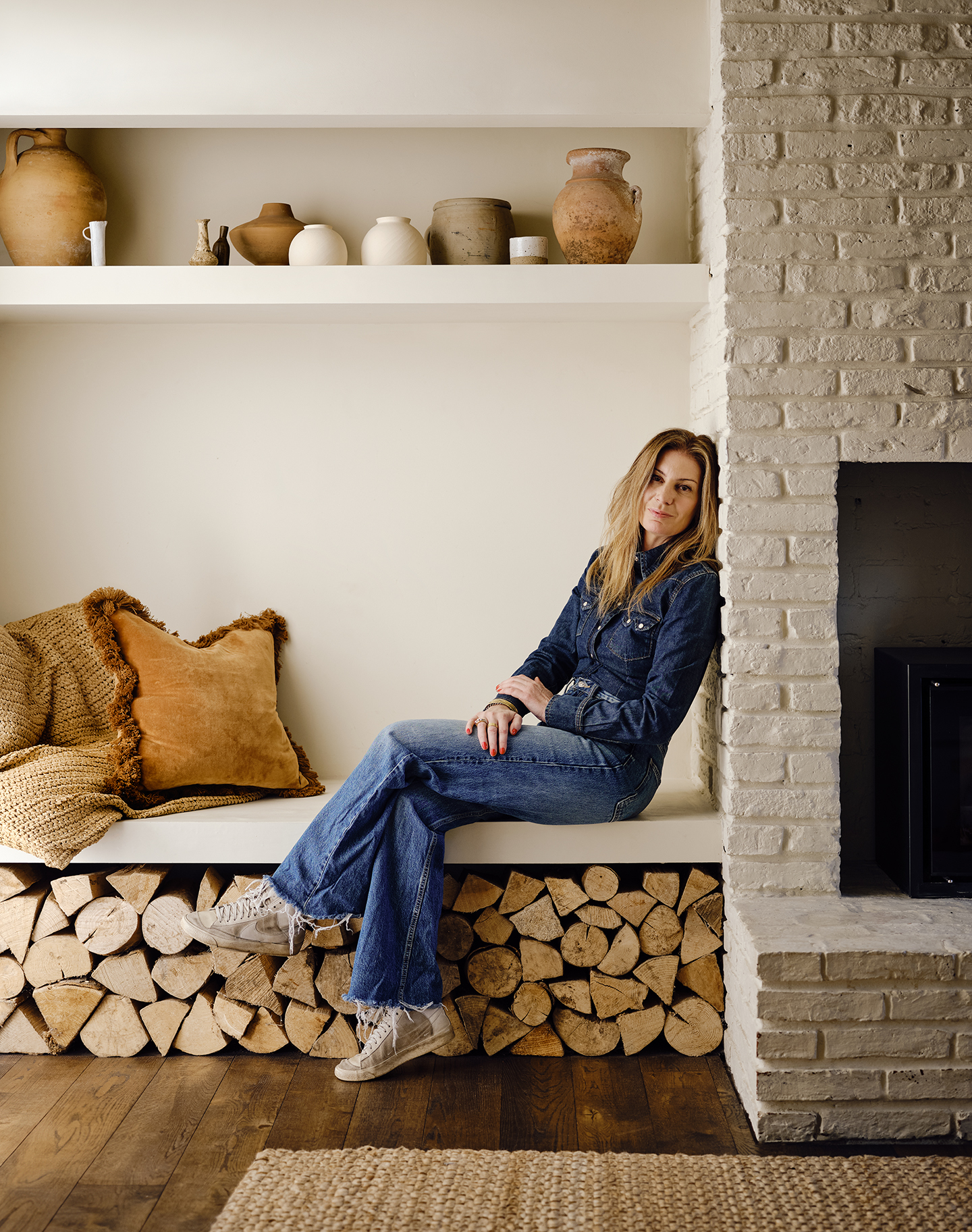 A woman sits on a built-in bench above a stack of wood logs cut for a fire.
