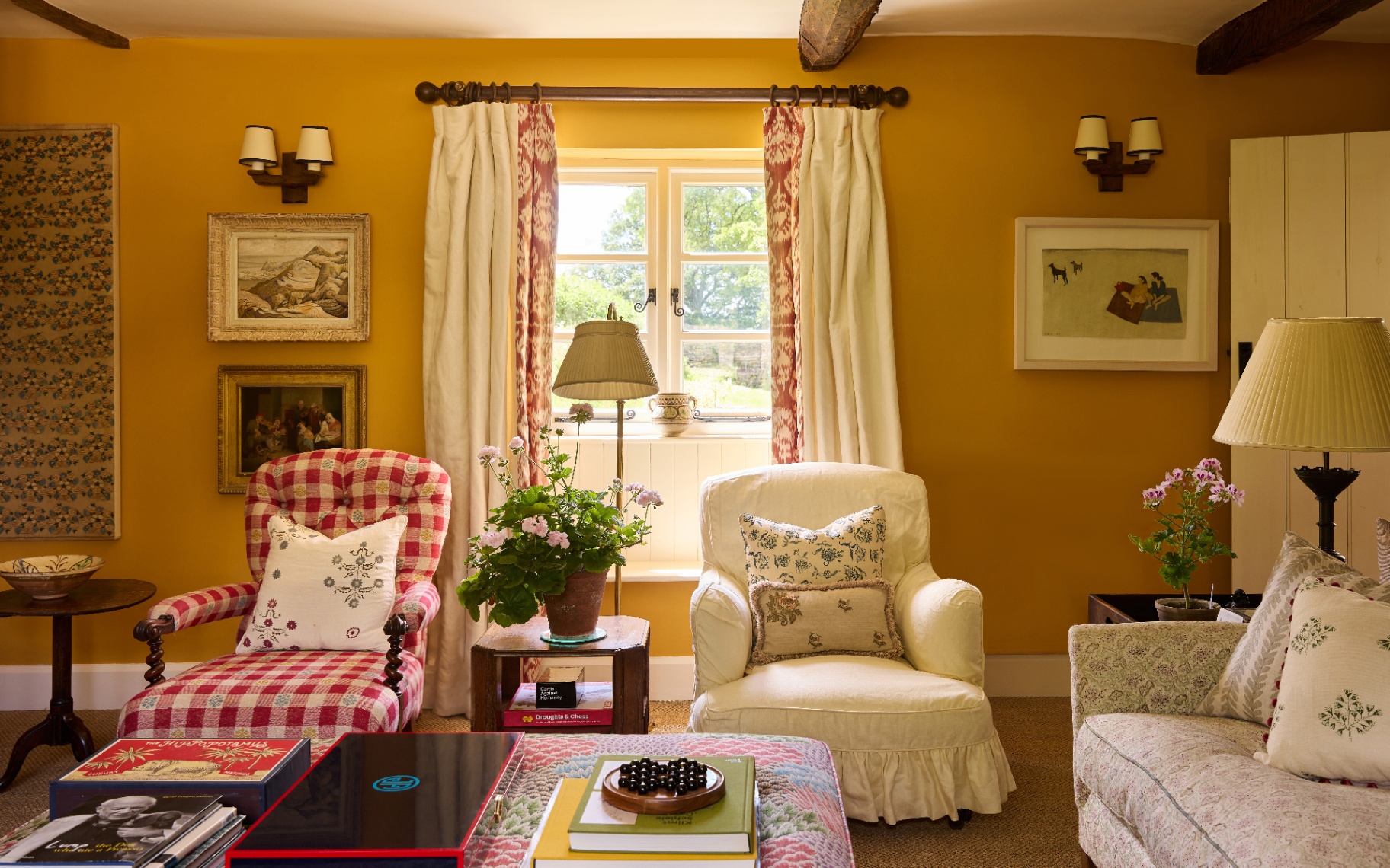 A warm, mustard yellow living room featuring a red and white gingham armchair, a soft cream armchair, floral cushions, wall sconces, and a window framed with pattern-edged curtains