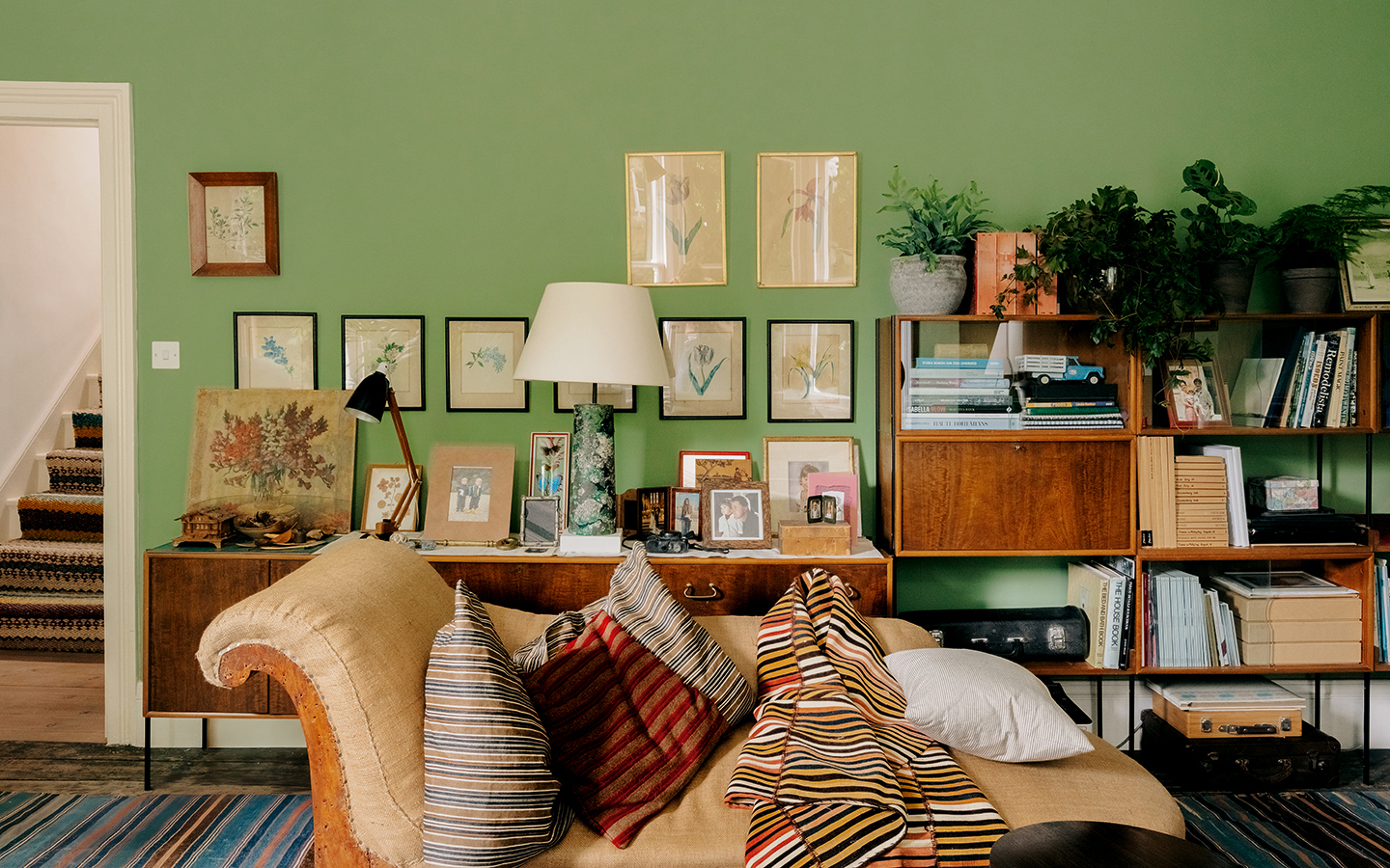 a green room with an antique chaise longue in the foreground and wooden cabinets and shelves topped with plants, books and trinkets run along the back wall, above hangs a selection of botanical artworks