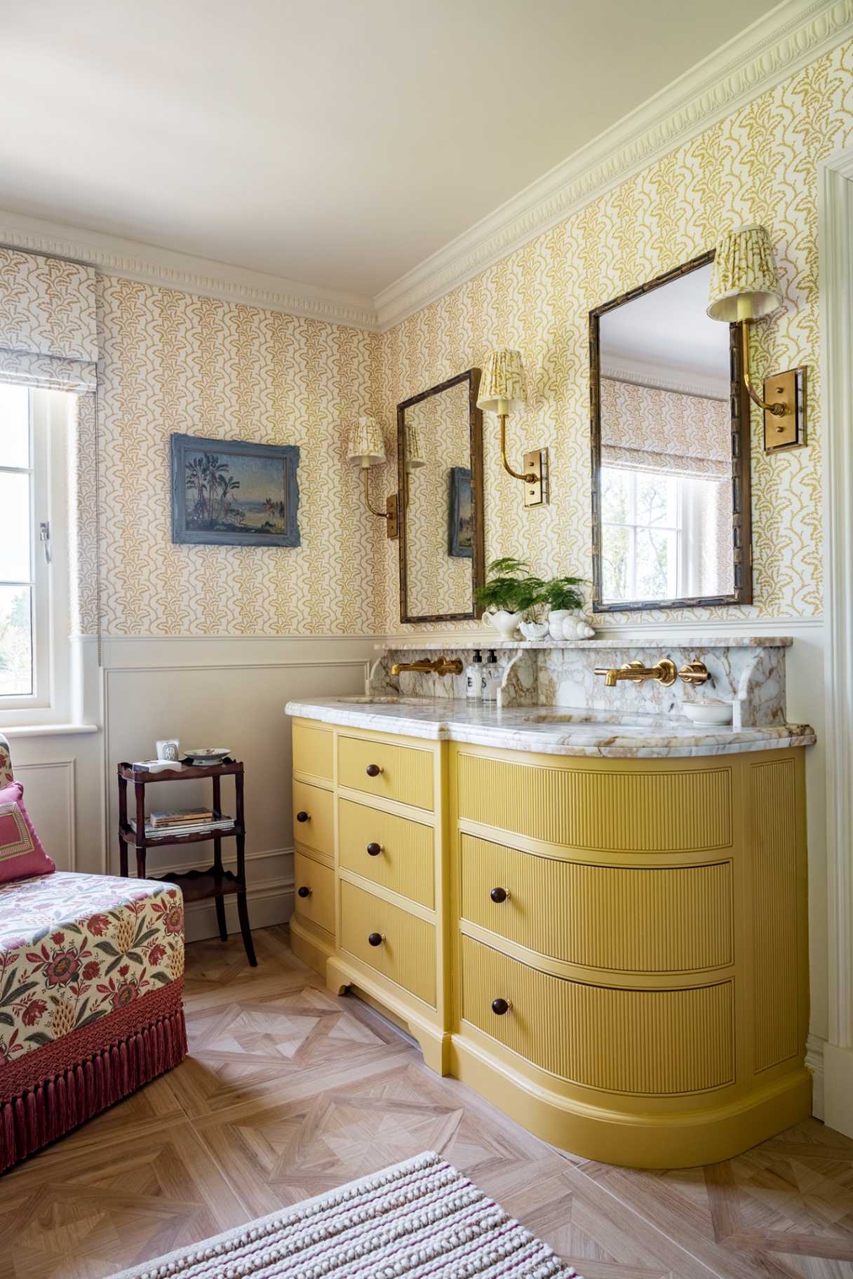A bathroom with a large double vanity painted yellow and with marble tops. Two mirrors and three wall lights hang above. The walls are papered in a patterned yellow and white striped paper