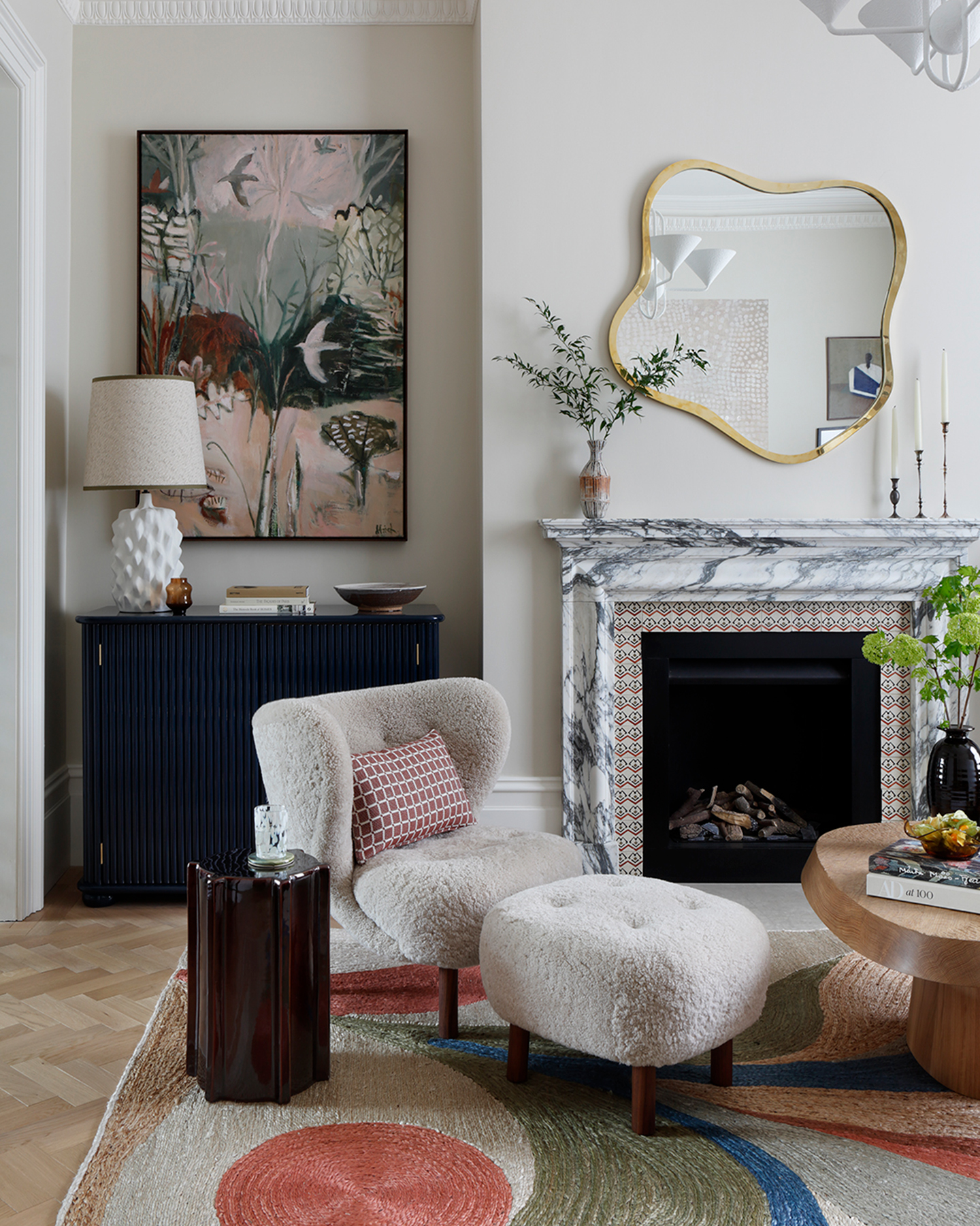 a living room with a large marble fireplace, a wiggly gold mirror above it, a borg armchair with matching footstool and an orange, green and blue swirl pattern rug