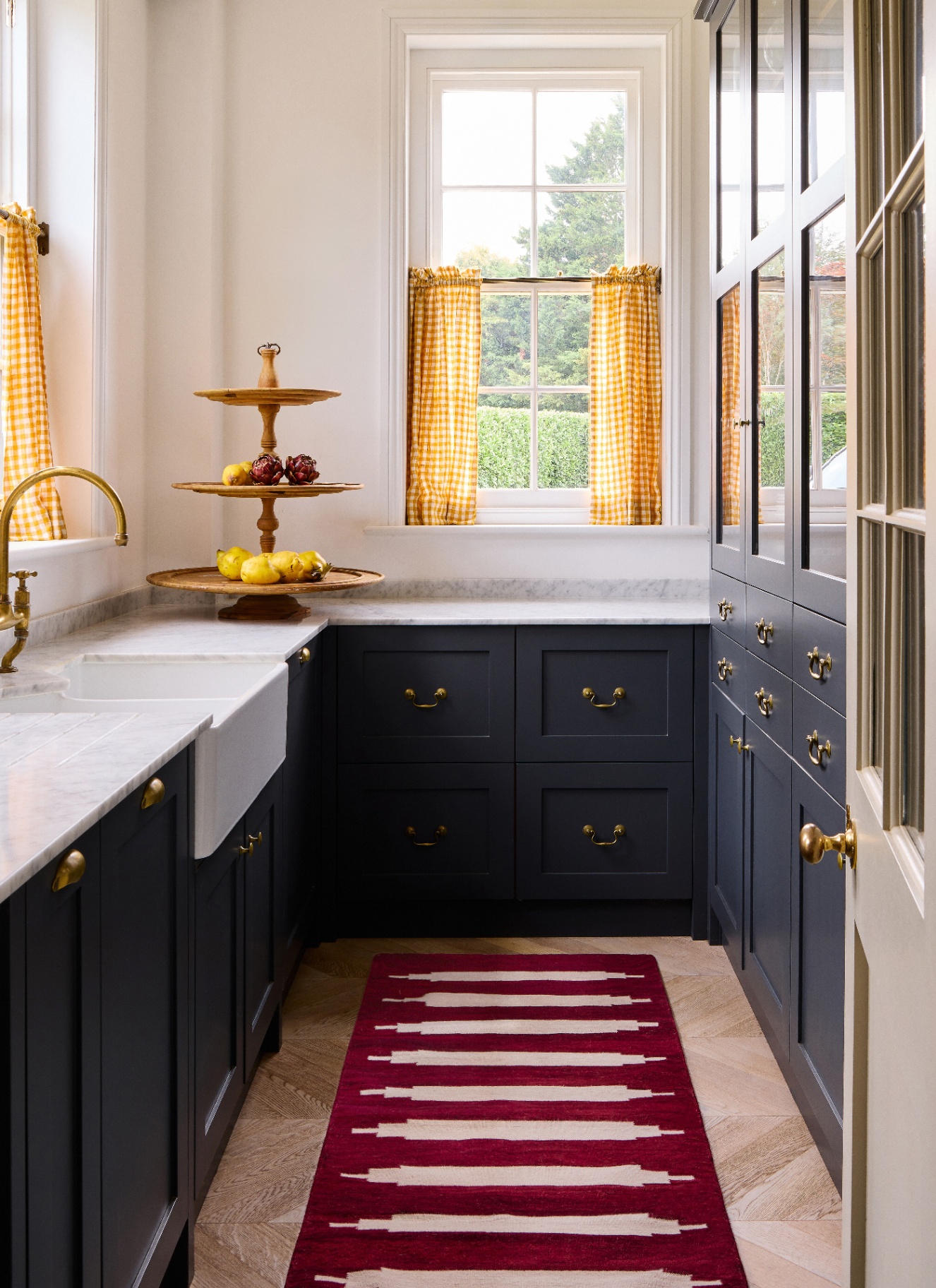 a narrow kitchen with dark navy cabinetry, a dark red and white runner, white marble tops and a window with yellow gingham cafe curtain