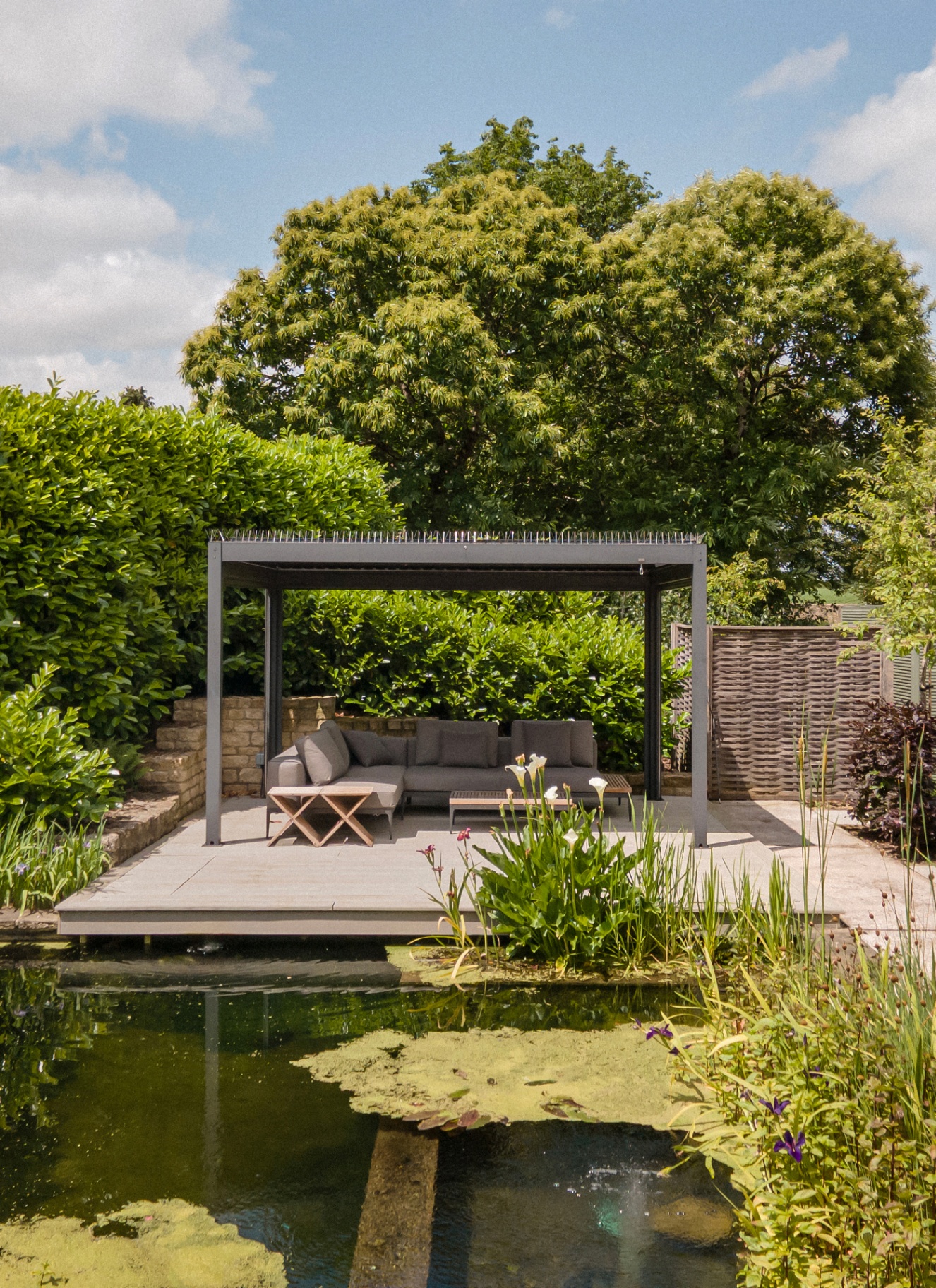 a large garden with a pond surrounded by greenery and a large grey pergola with furniture beneath it