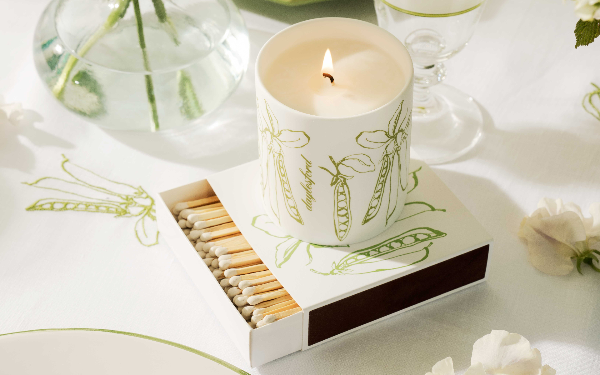a table with a lit ceramic candle with pea illustrations on top of a matching matchbox on a white table cloth