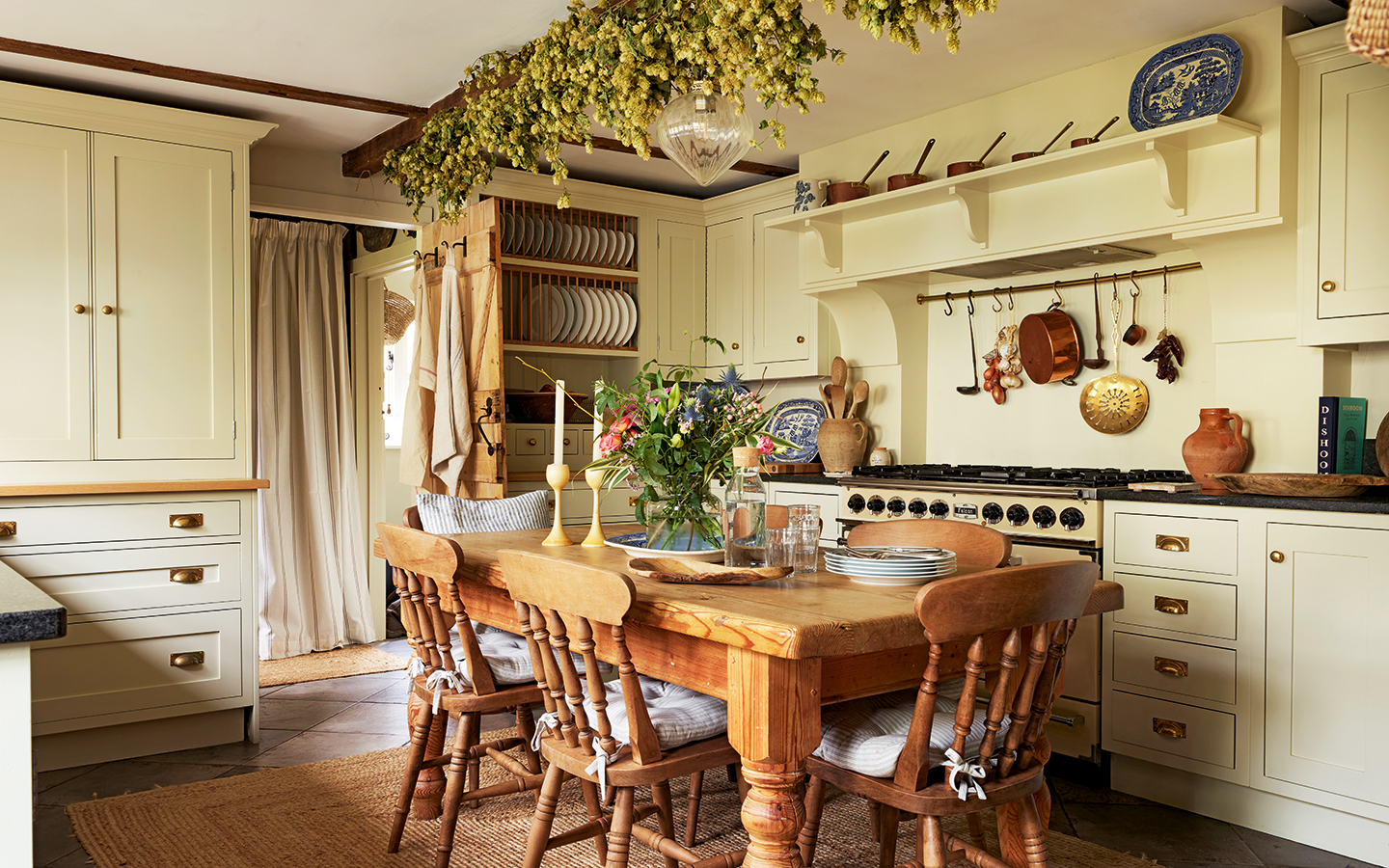 A country kitchen with wood table and chairs in the centre on a jute rug atop a stone floor. Cream cabinets surround the seating area plus a cream cooker. Hops decorate a beam running across the ceiling above the table