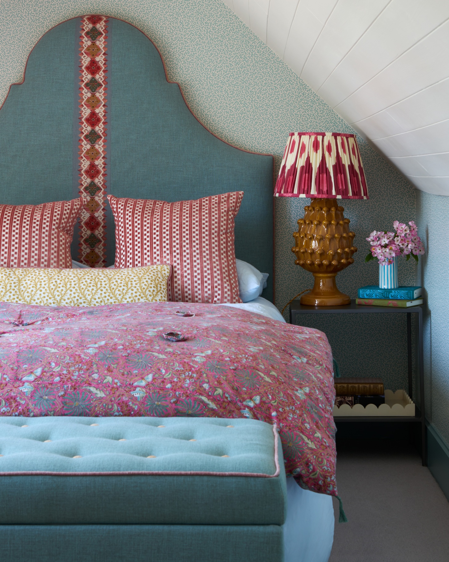 a small bedroom with a bed with a large blue upholstered headboard, two red patterned cushions, one yellow pattenred cushion a red floral bed spread, ablue storage ottoman at the foot of the bed and a large ochre lamp with a red ikat shade on a table