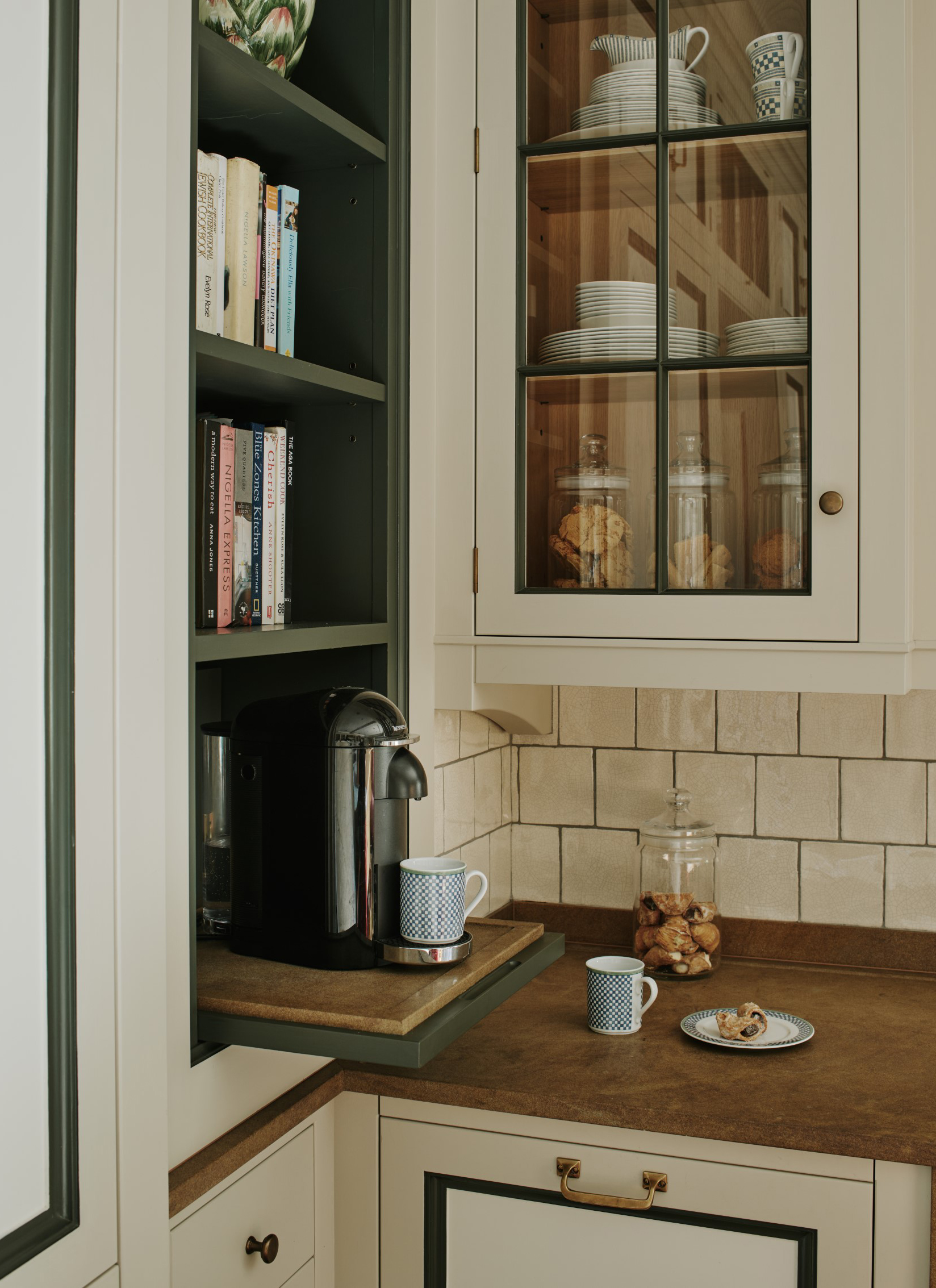 Compact kitchen coffee nook featuring a black pod coffee machine resting on an extended wooden pull-out shelf, situated beneath dark green cookbook shelving and a glazed wall cabinet
