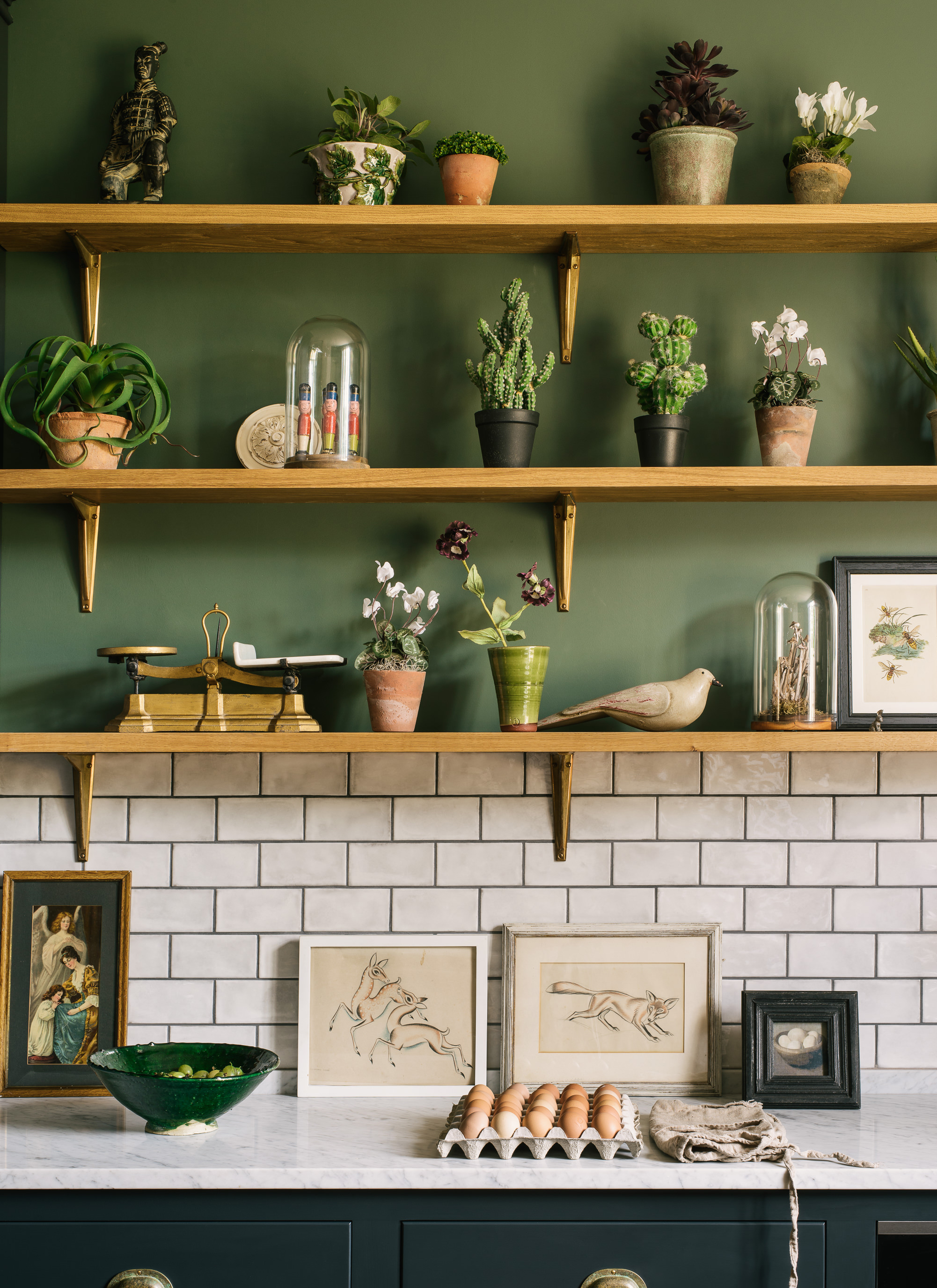 a close up of a kitchen area with dark blue cabinets, white marble counters, a white tiled splashback, green walls and 3 open wooden shelves with plants on