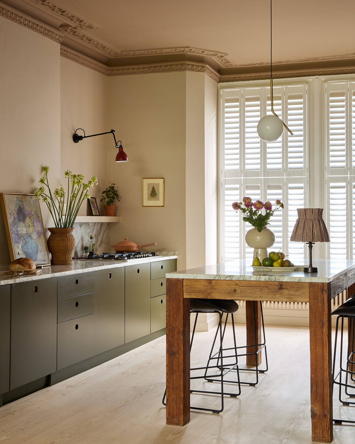 a kitchen diner with cream walls and a beige ceiling, a wooden table with a grey marble top and a small pleated lamp, dark green kitchen cabinets with a pink frying pan on the hob and a large rattan vase with tall green flowers
