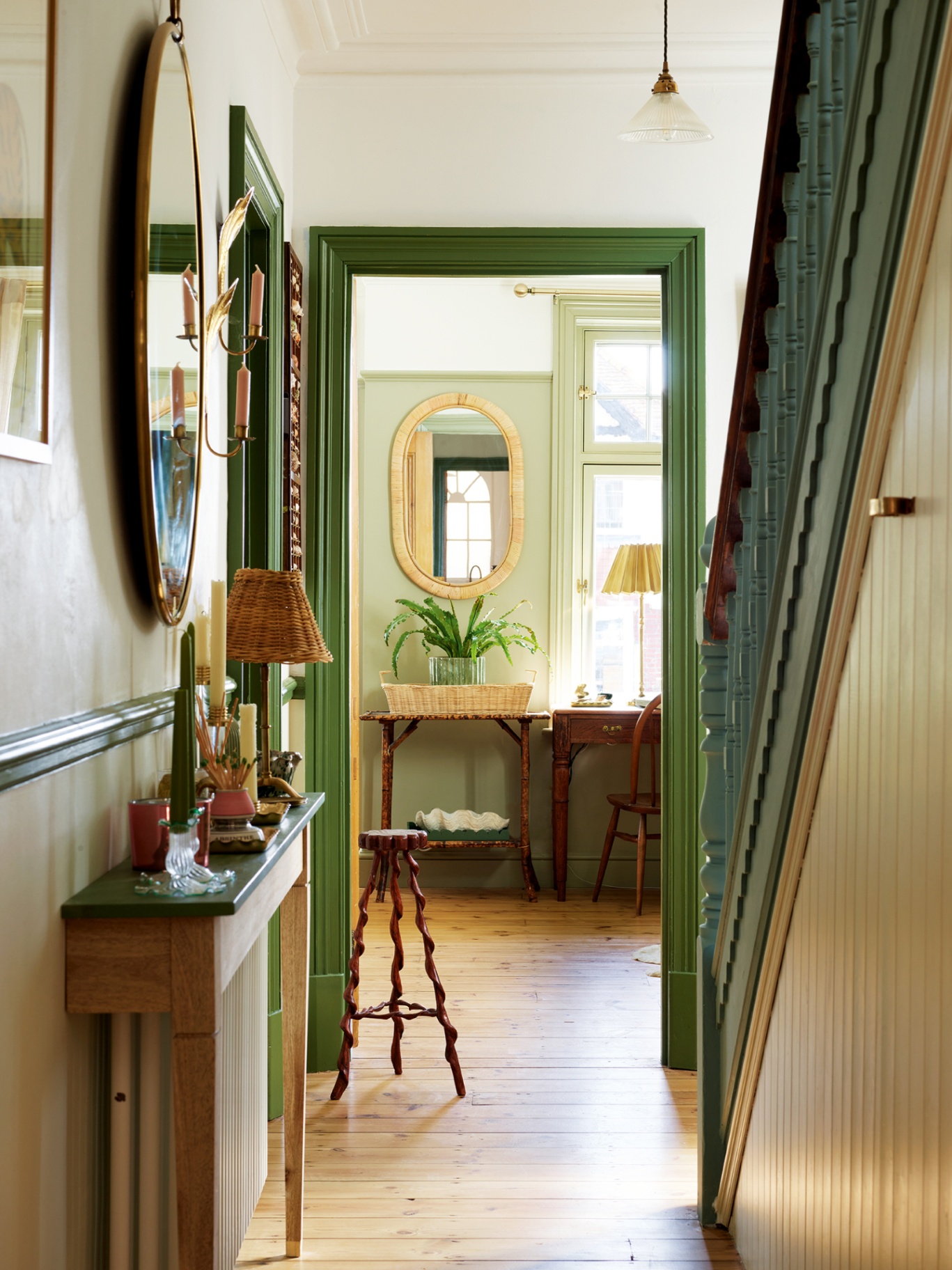 The narrow hallway of an Edwardian house with the stairs to the right. The walls are painted in white, the woodwork in green and the stairway in blue. A small console sits to the left topped with a table lamp, candlesticks and matches