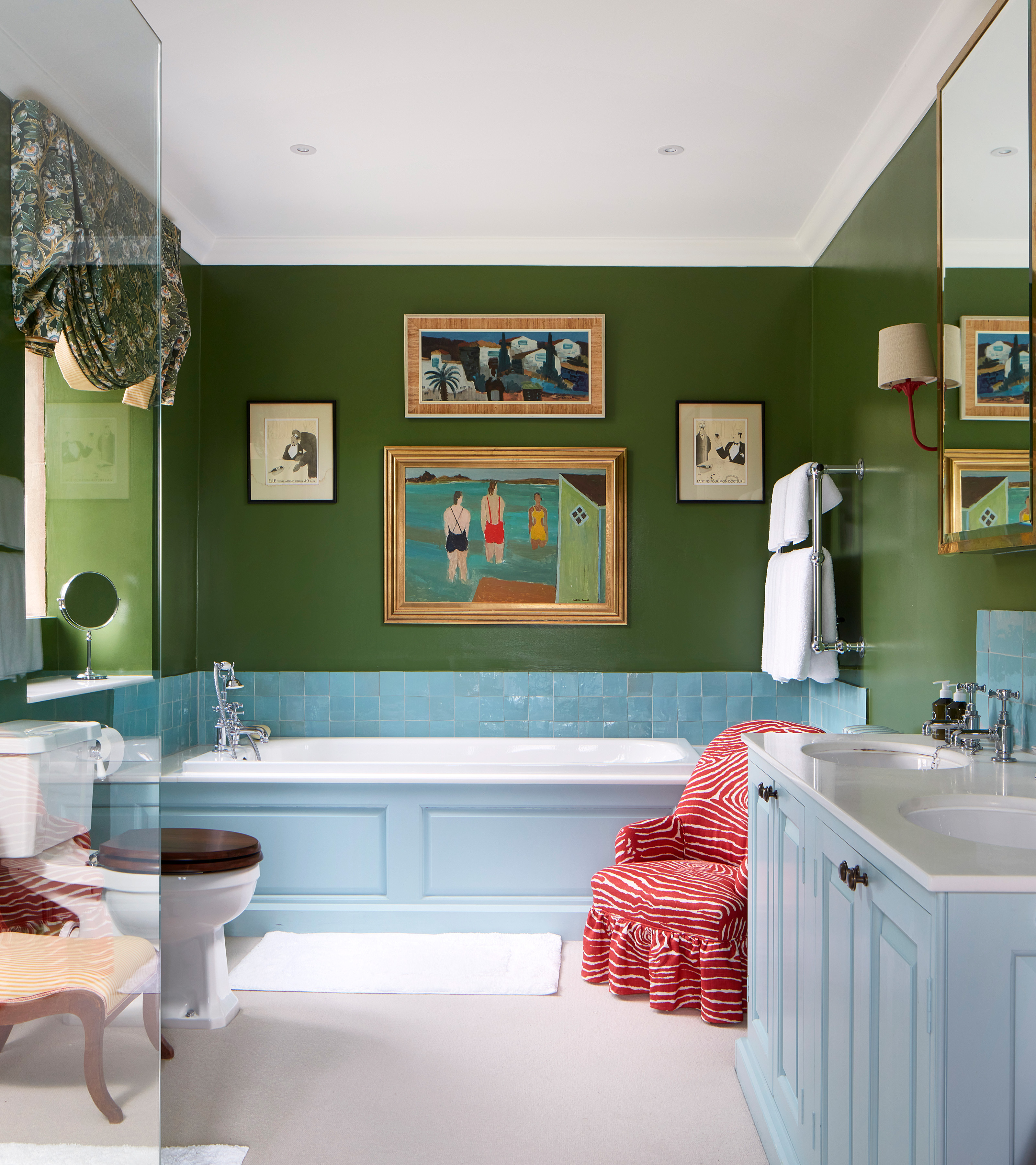 a bathroom with forest green walls, pale blue vanity and bath with a red patterend chair next to it and a gallery wall above the bath