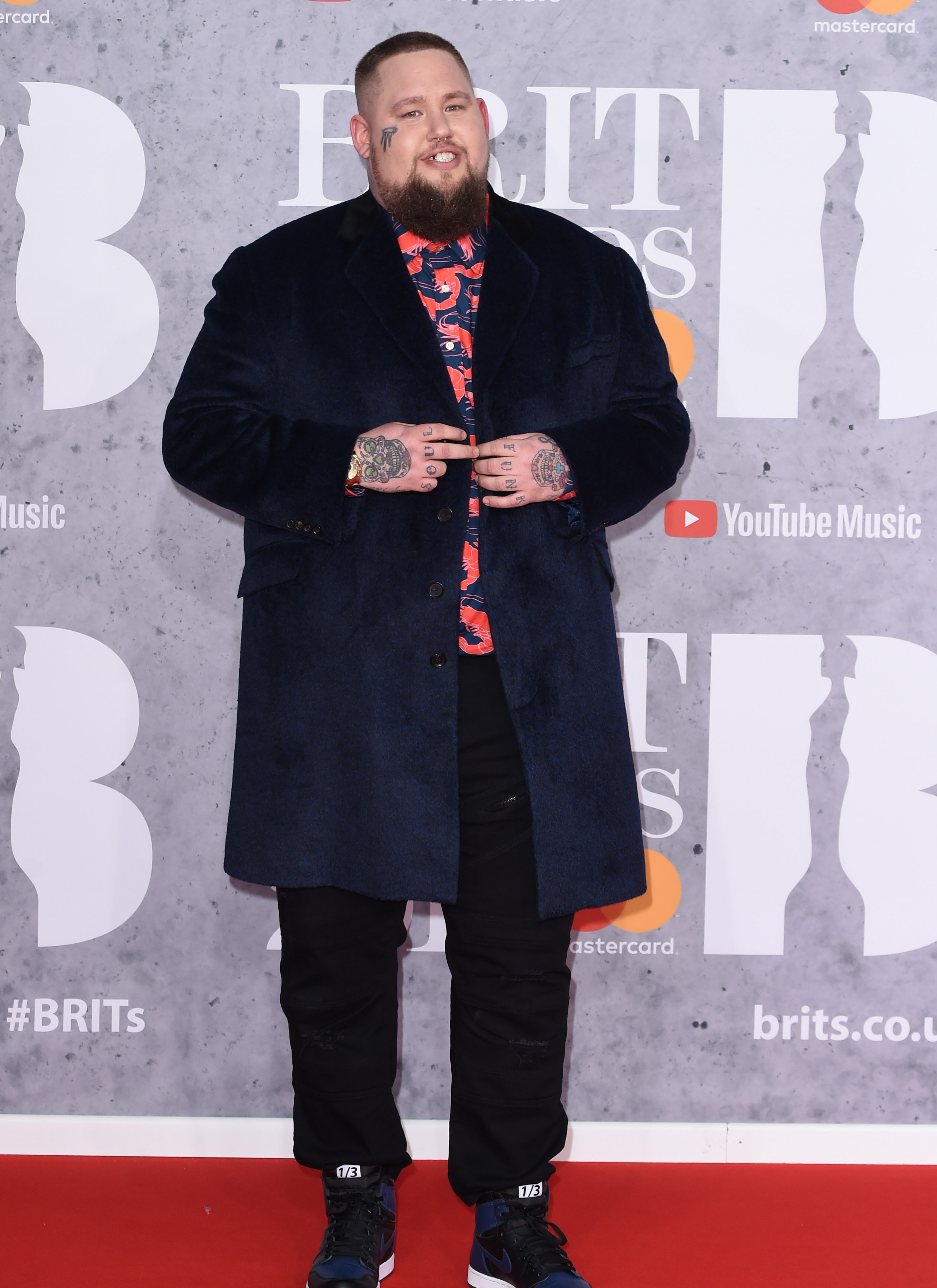 rag n bone man on the brit awards red carpet wearing a long black coat, black trousers, an orange patterned shirt and black trainers