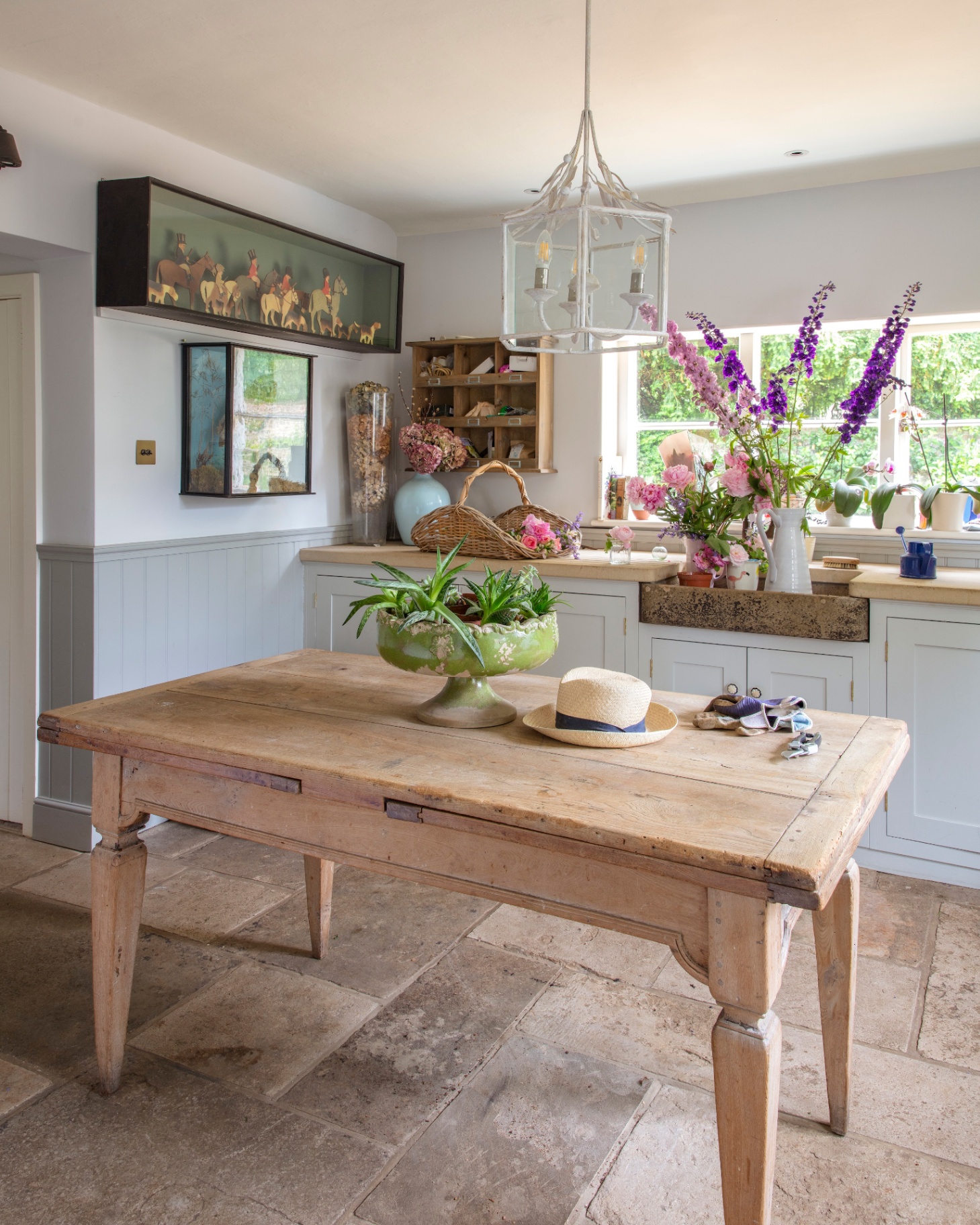 a kitchen with a large farmhouse table in the centre, flagstone flooring, pale blue cabinetry with vase of purple flowers