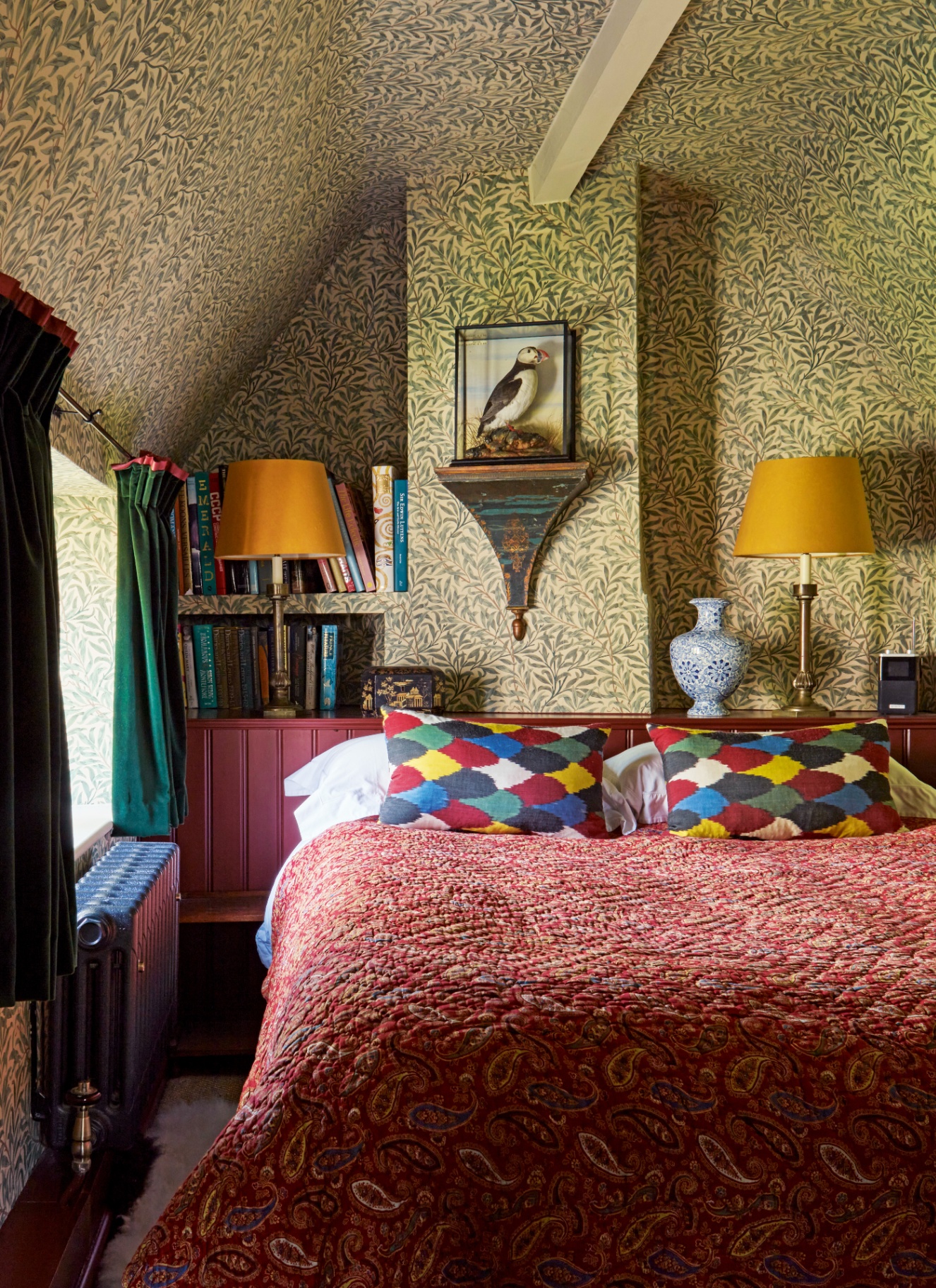 a bedroom with sloped ceilings and leaf wallpaper, a recessed bookcase with colourful books, a bed with a red patterend bedspread and a puffin ornament on the wall
