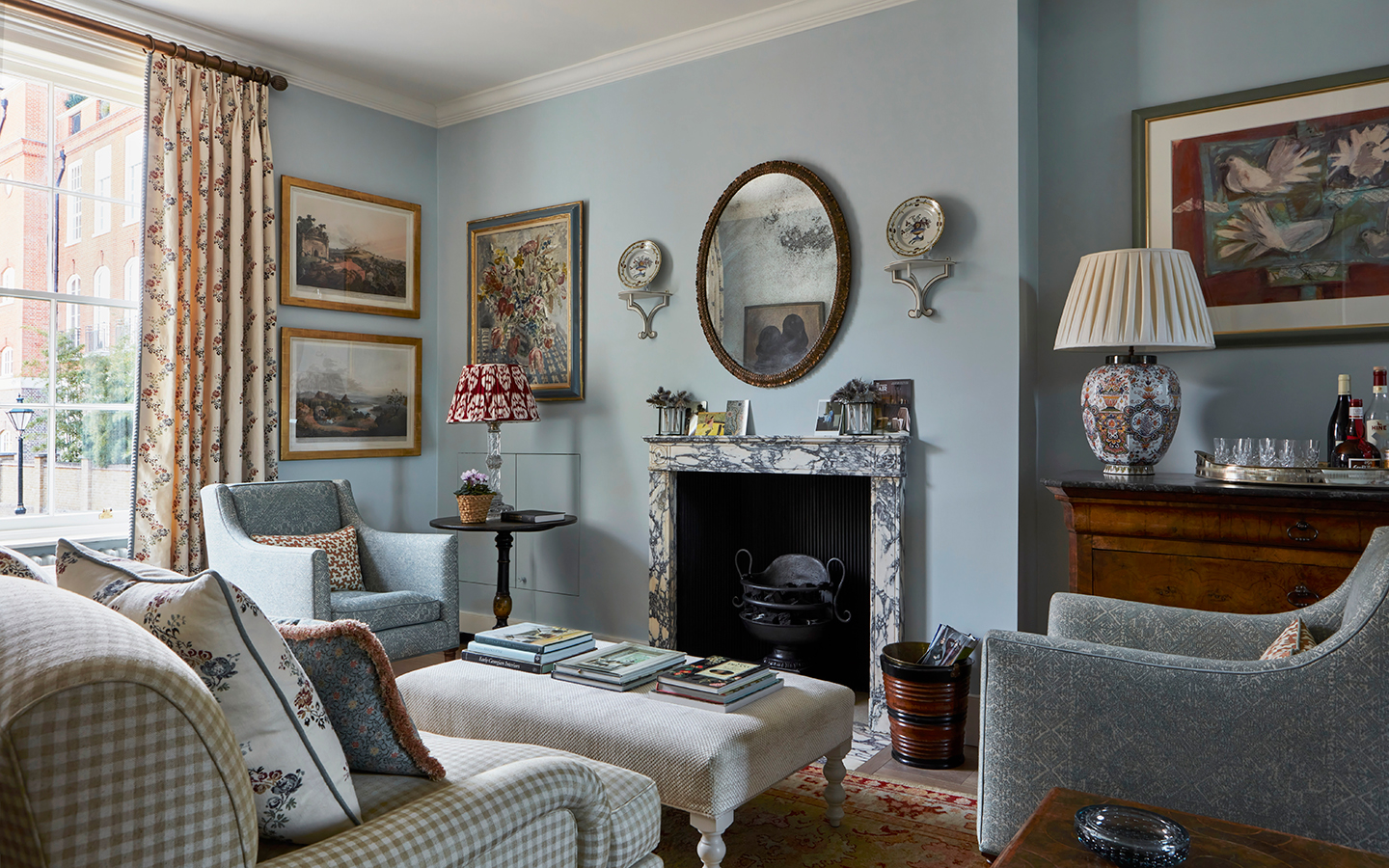 a blue living room with two matching blue armchairs and a check sofa surrounding a cream ottoman. There is a marble fireplace and antique art, plates and a circular mirror hanging on the walls