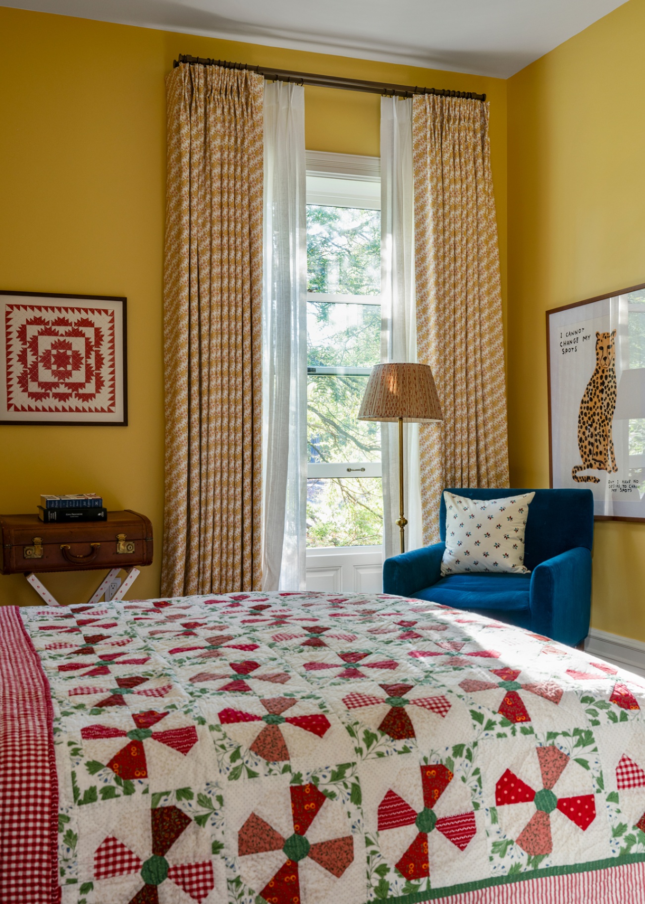 A rich yellow guest bedroom with geometric print curtains dressing a window with a floor lamp and blue armchair in front. In the foreground is a double bed dressed with a red and green patchwork quilt