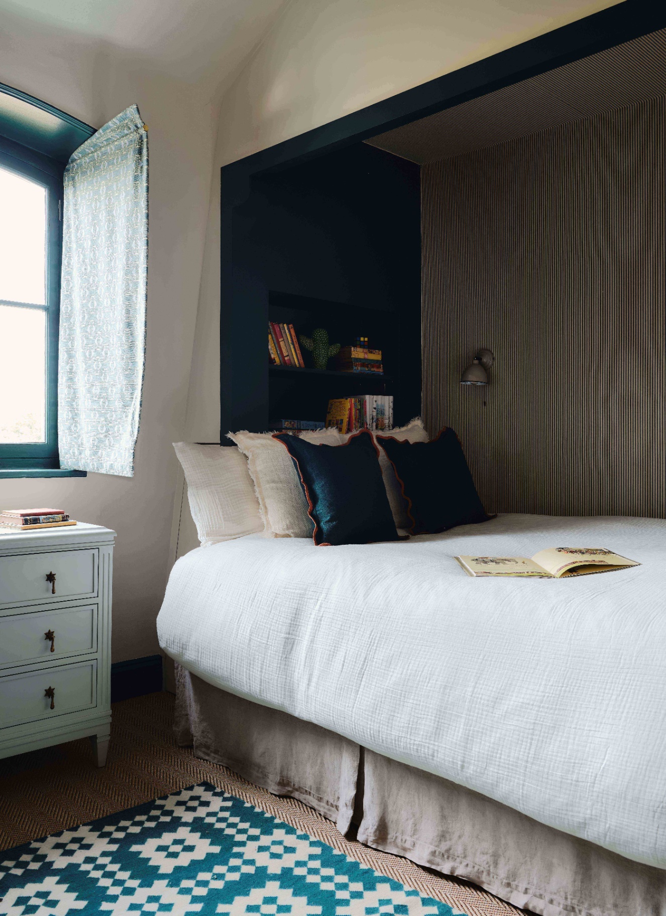 a bed nook with white bedding and striped wallpaper behind the bed, a built in shelf above the headboard and a blue and white geometric rug