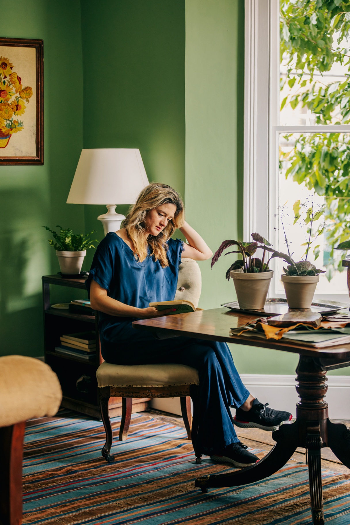 interior designer holly howe sits at a square wooden table in a green painted room with a striped blue and brown rug underfoot