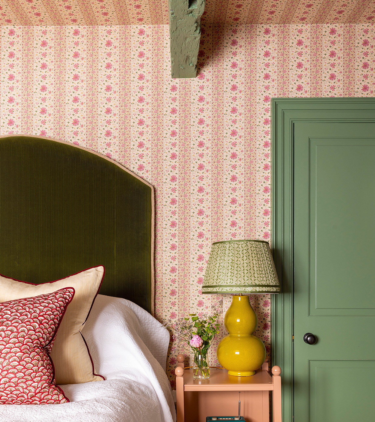 A bedroom with pink ditsy floral wallpaper, a green painted door, a peach bedside table with a yellow table lamp and a bed with white linen and a green headboard