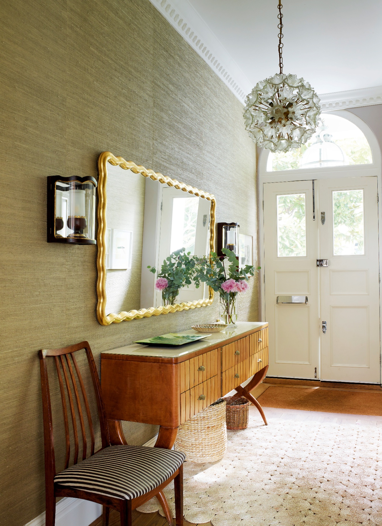 a hallway with green walls, a large gold wavy mirror, a wooden console and an orb pendant light