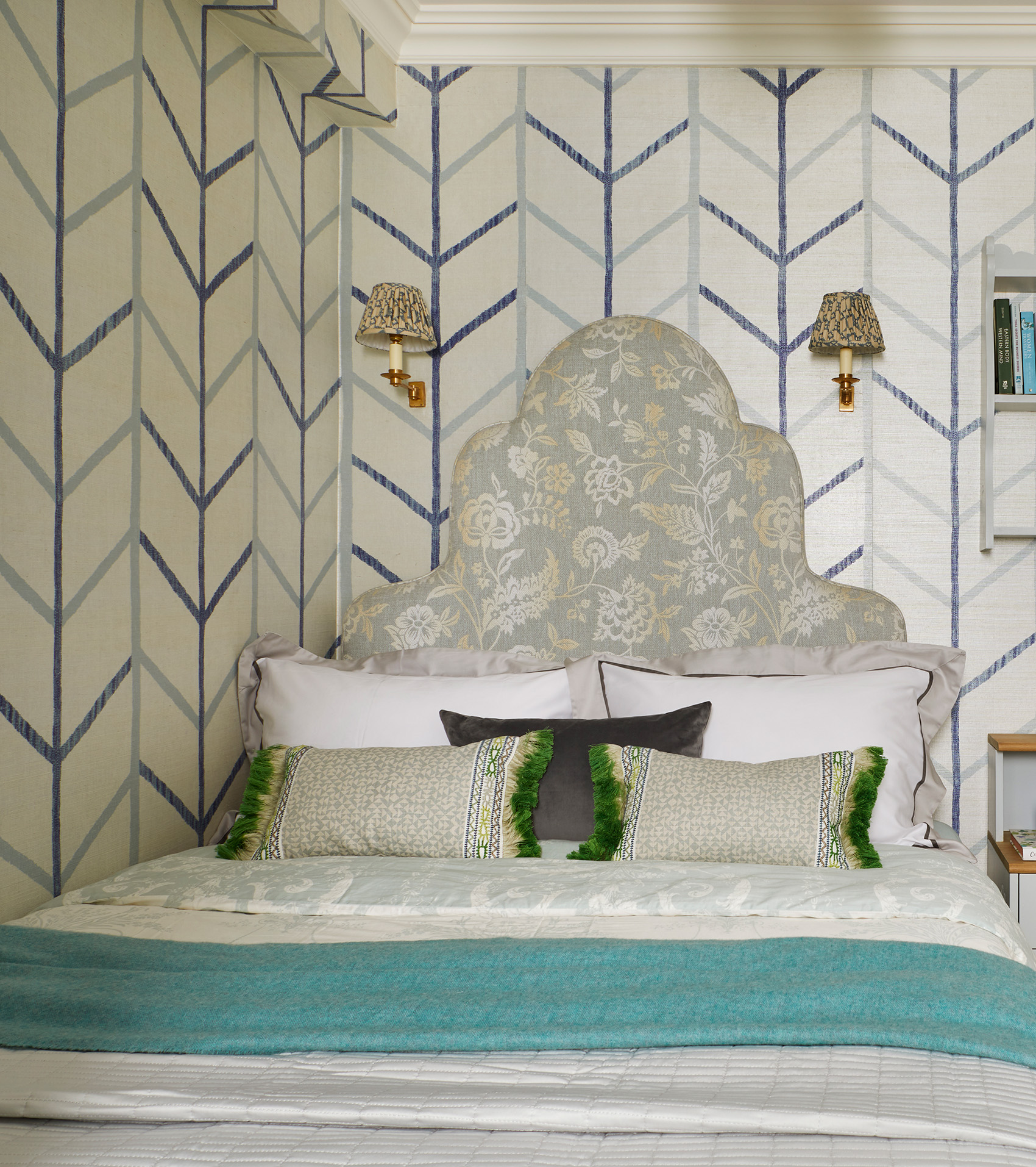 a bedroom with a blue and white striped wallpaper, two wall sconces with pleated shades and a small double bed with a green floral headboard and white bedding