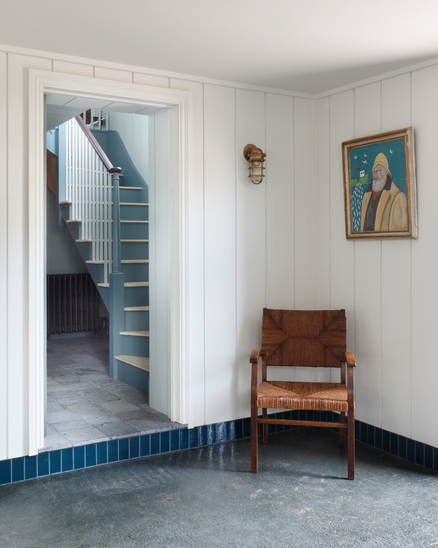 a hallway with white paneled walls, a nautical inspired brass wall light, a painting of a fisherman on the wall and a brown wooden armchair