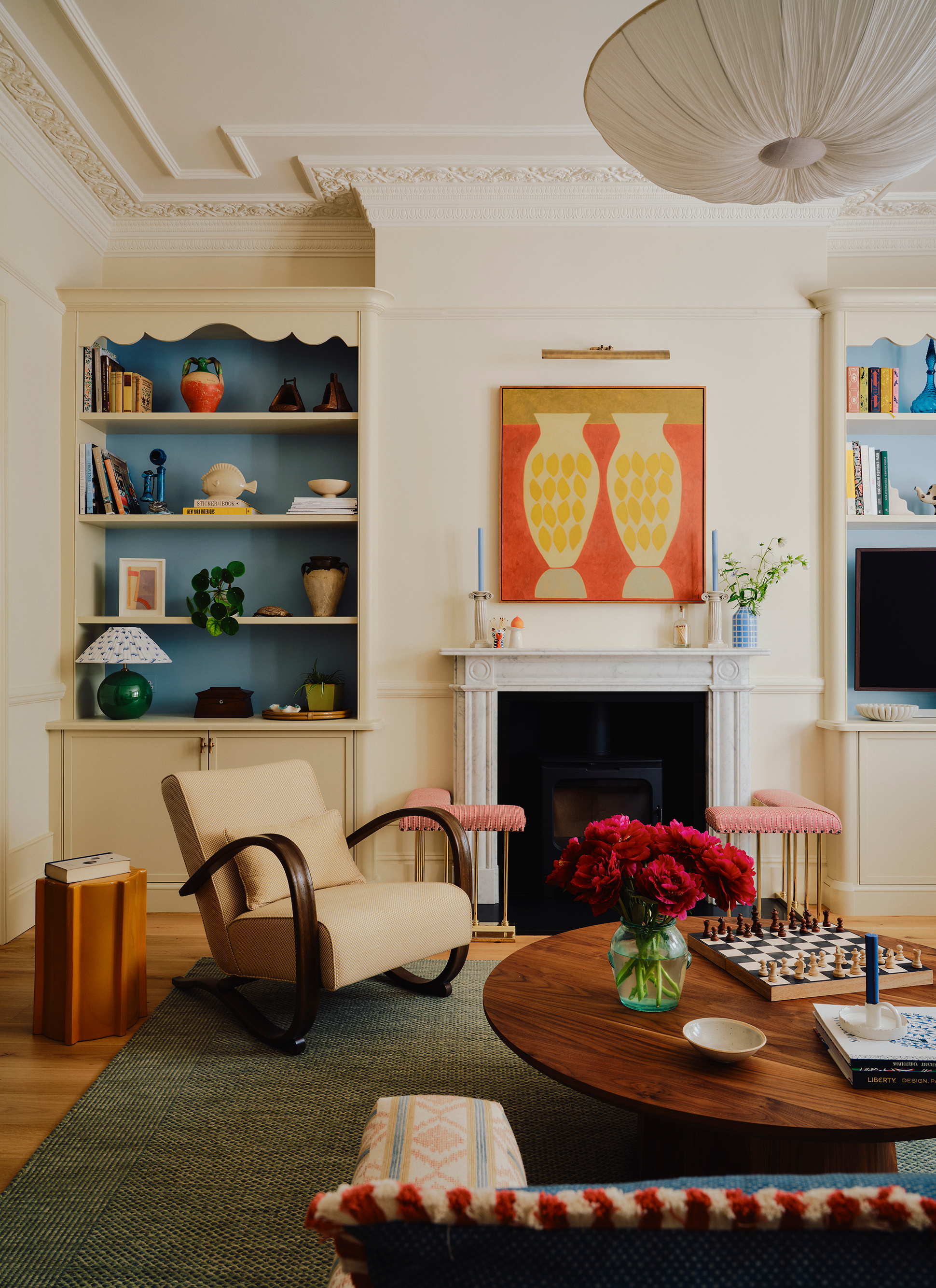 a living room with cream walls, a large bookcase, a fireplace with an orange and yellow artwork above it and a cream armchair with a wooden round coffee table