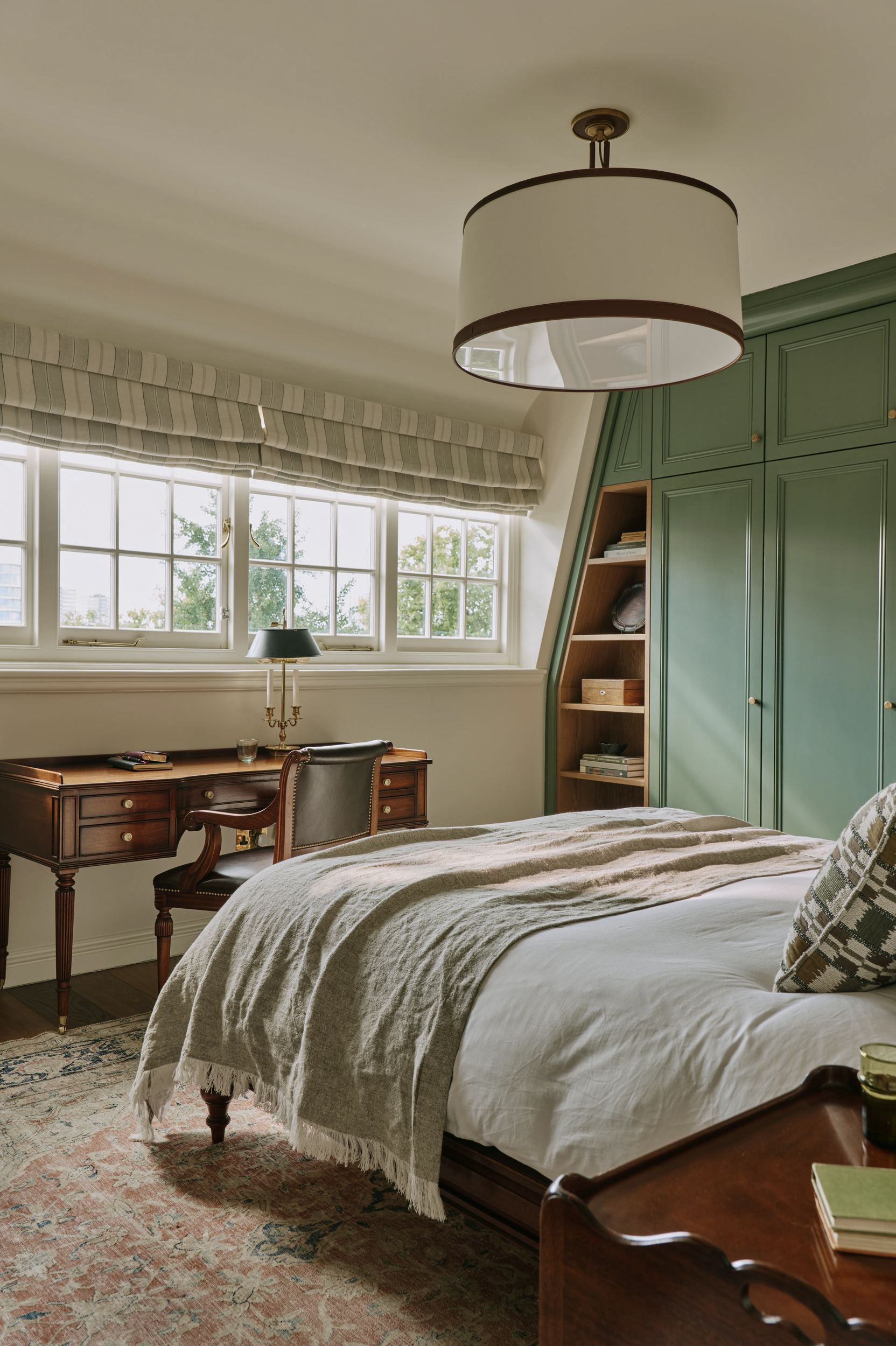 Angled shot of a bedroom with a double bed in the centre, a green built-in wardrobe and shelving unit and a wooden desk and chair underneath a row of windows dressed with striped roman blinds. A traditional-style rug covers the floor 