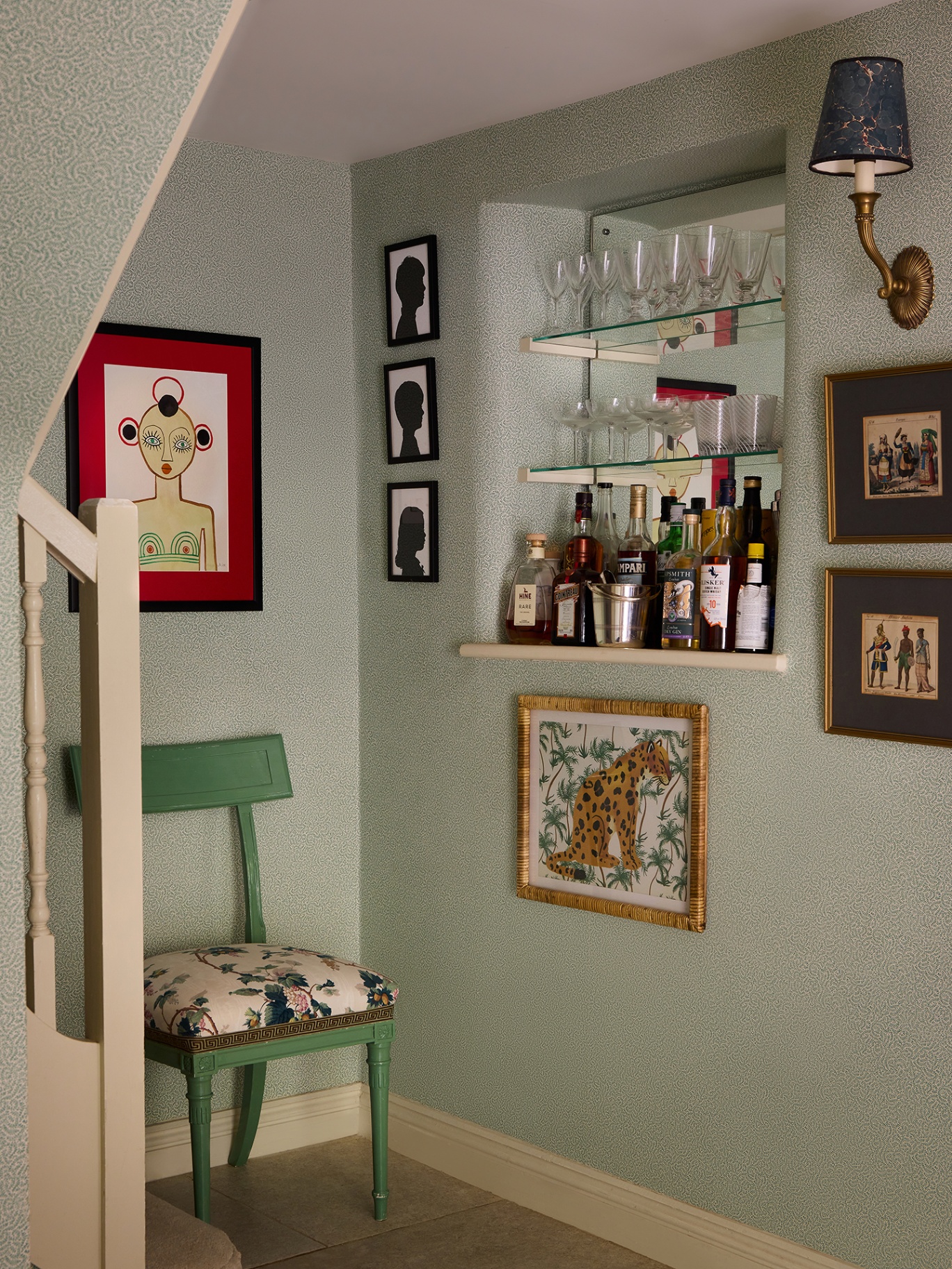 A hallway corner lined with smallprint wallpaper. In a mirrored nook shelves hold glassware and bottles. A green chair is placed in the corner and art and a wall light is hung on the walls