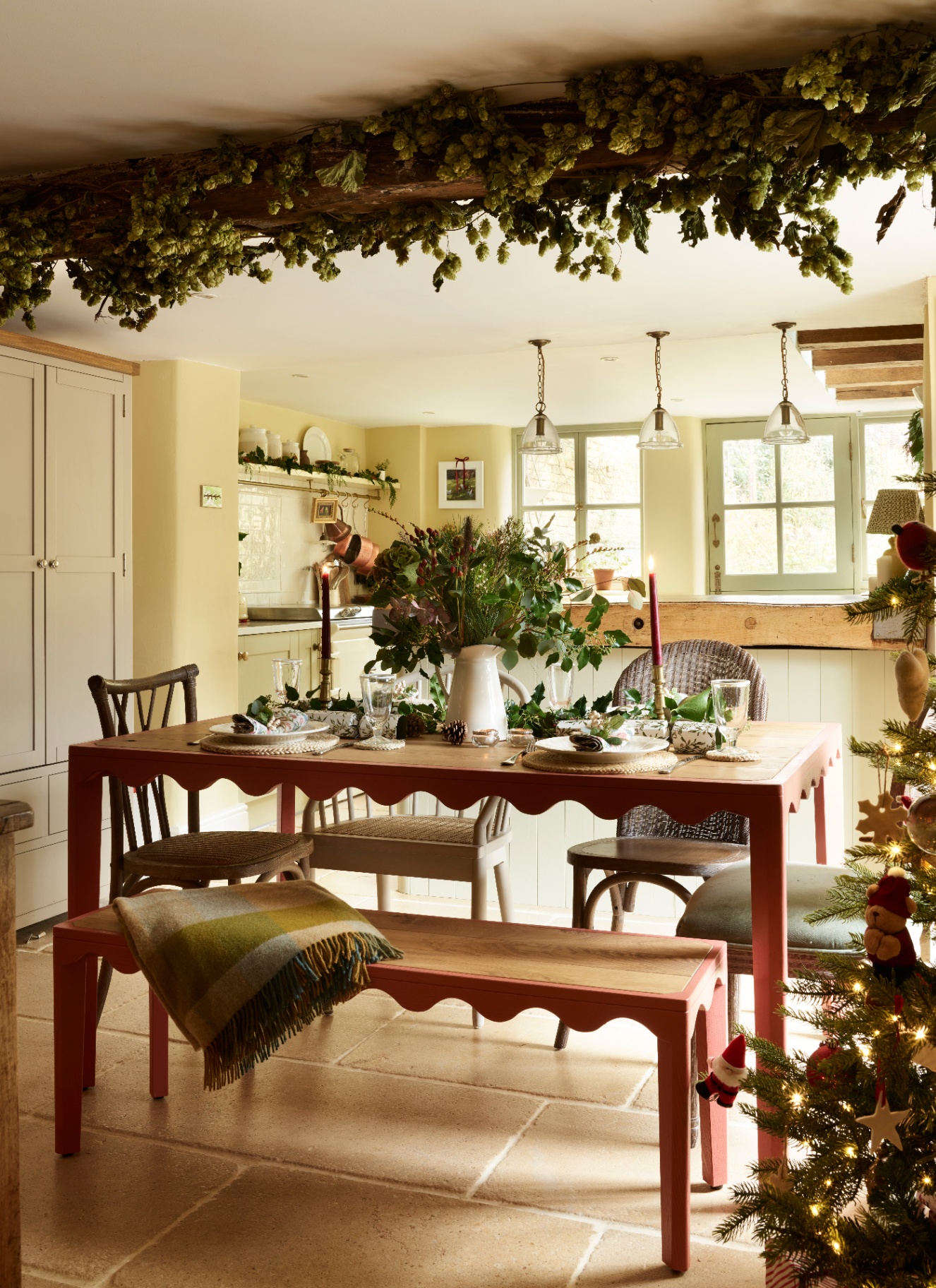 A crimson scallop-edged dining table set for Christmas with greenery, candles, and crackers under a beam strung with dried hops