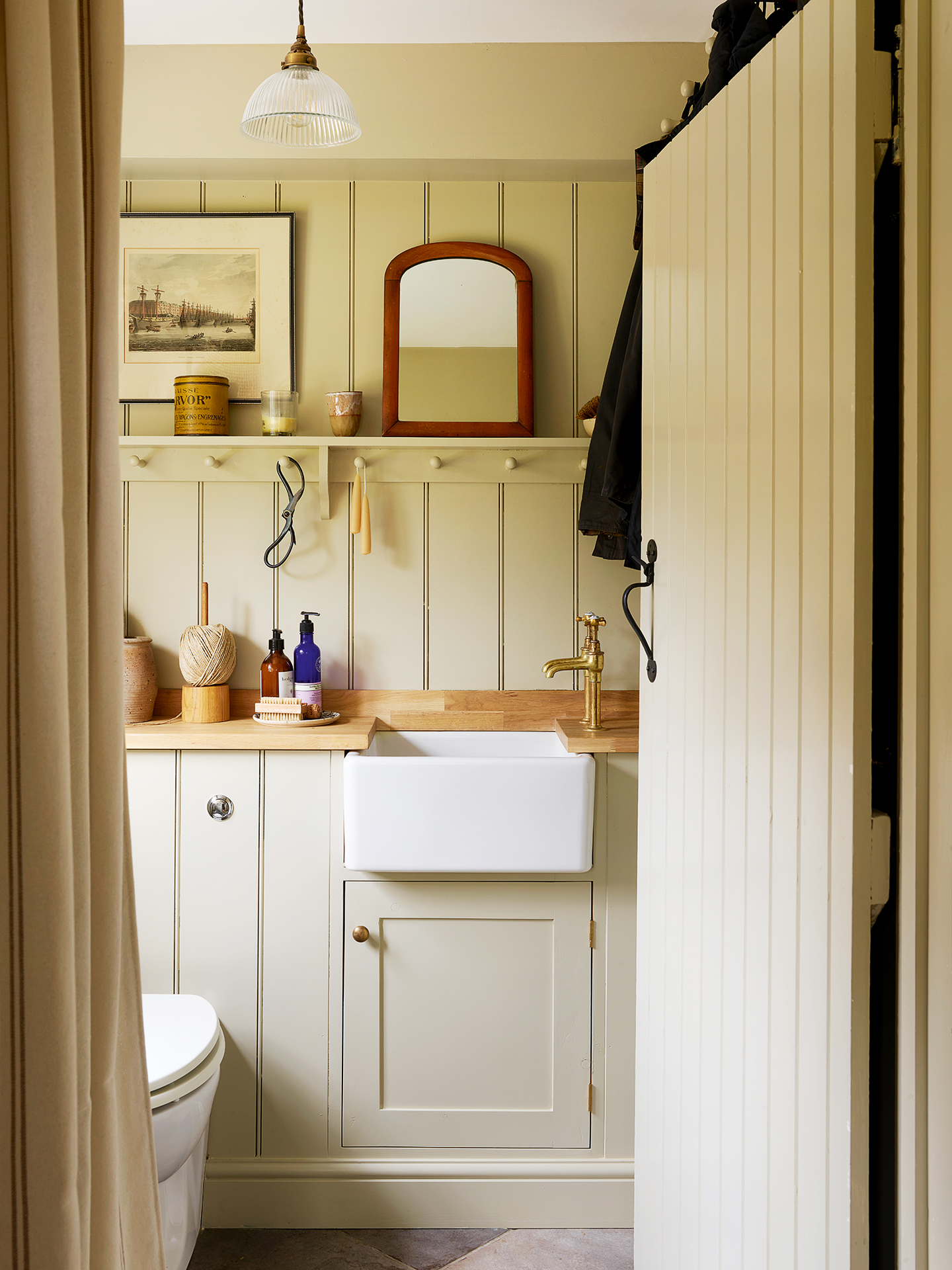 Country-style cloakroom with tongue-and-groove walls. A built-in vanity unit holds a sink, a toilet sits to the left and above is an open shelf with hooks. Toiletries, candles and trinkets are arranged on the unit worktop and shelf