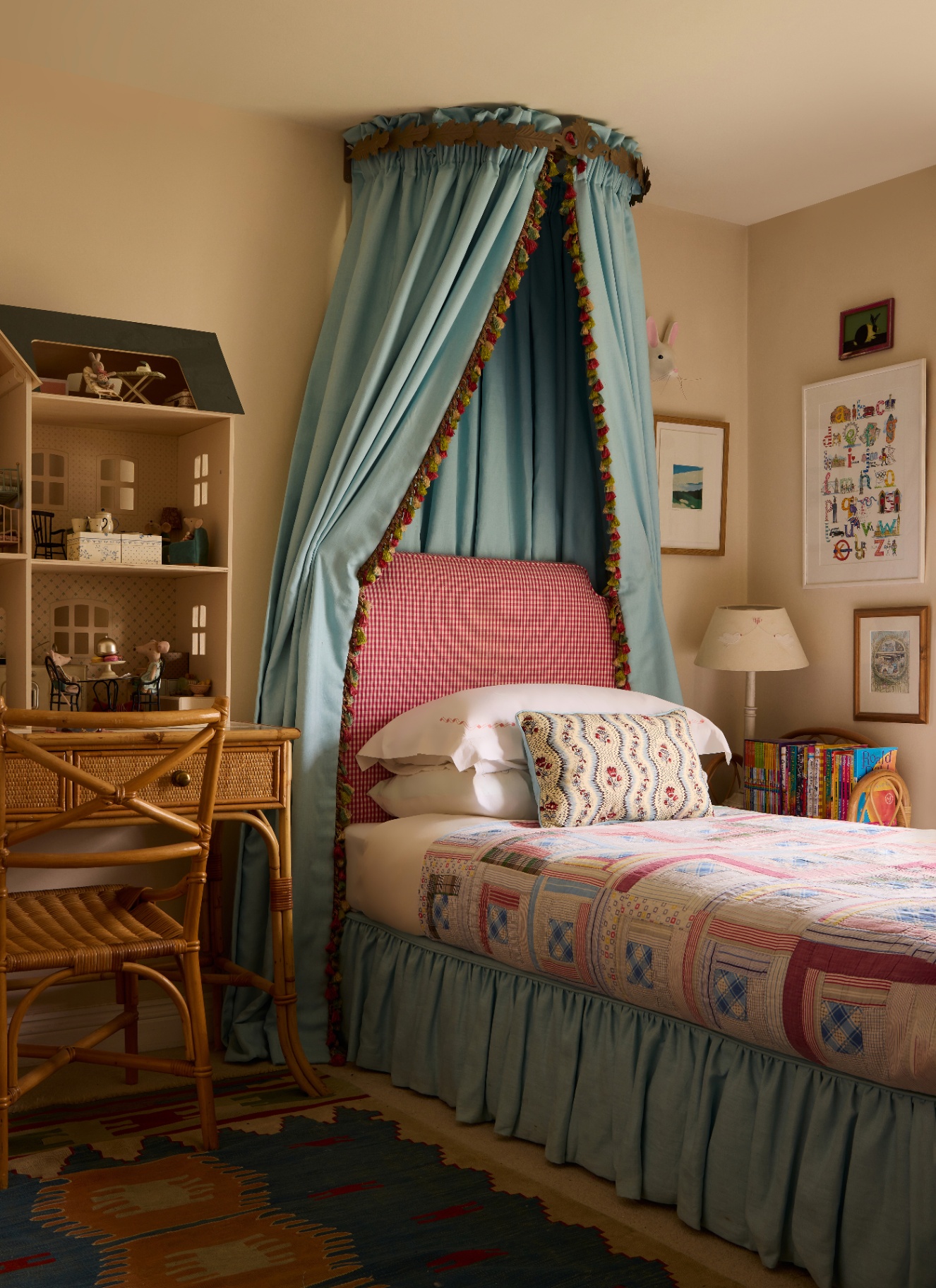 a children's bedroom with a single bed with a blue valance and a blue canopy witha pom pom trim and a rattan desk and chair with a doll's house