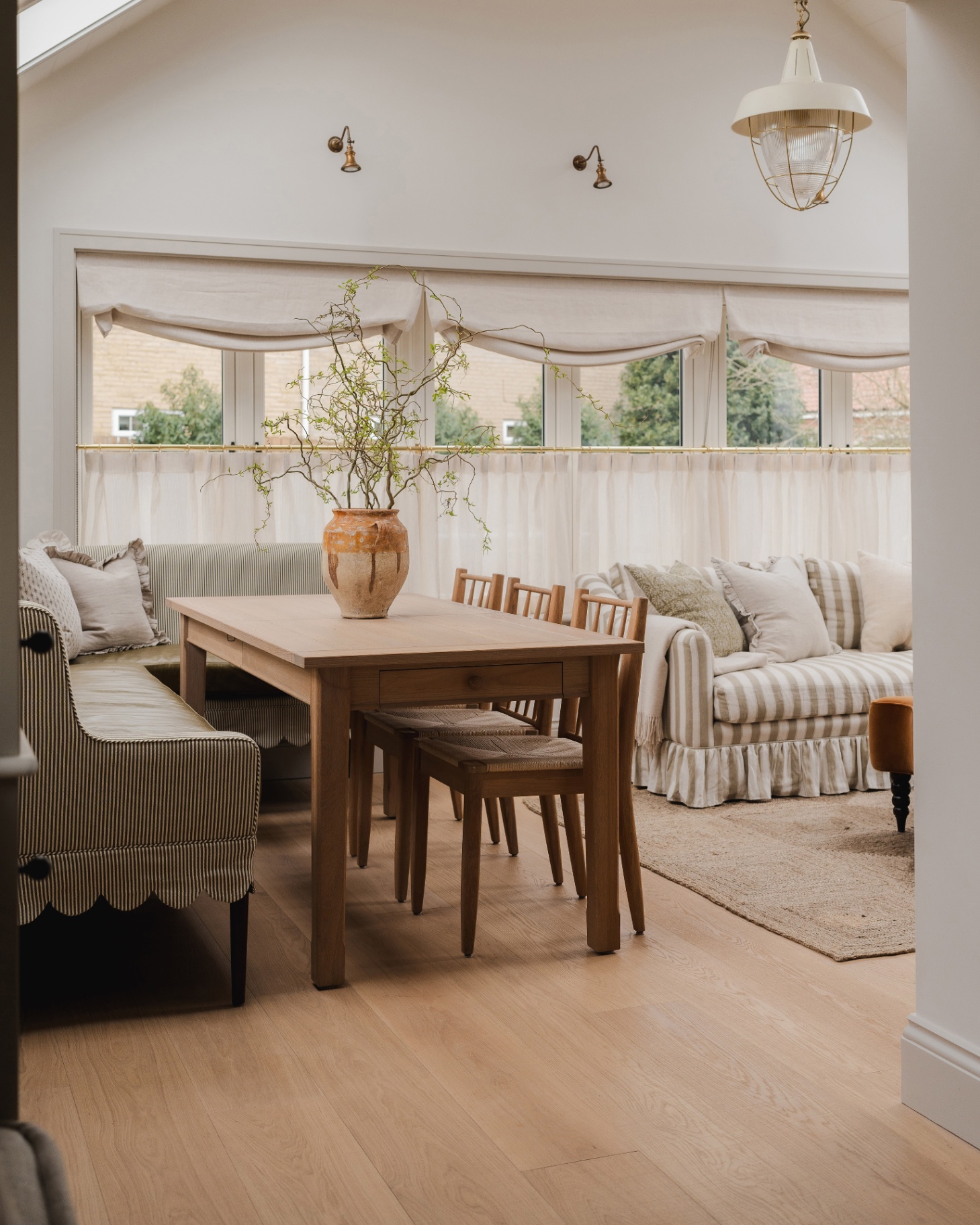 a dining room with a pale wooden floor, a wooden dining table with 3 wooden chairs and an L shape striped bench, a white and cream striped skirted sofa and a jute rug