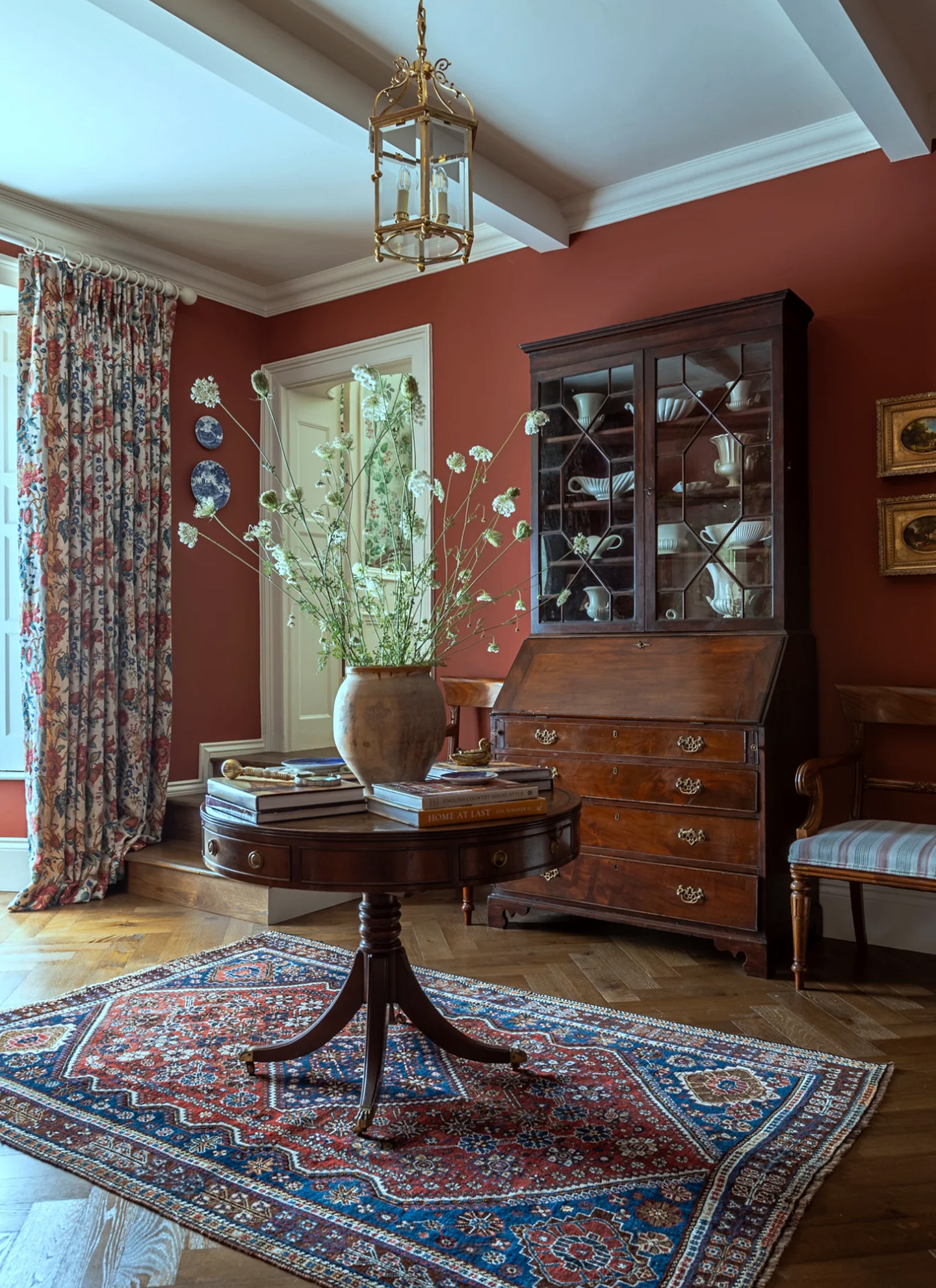 A tall, airy arrangement of delicate white seedheads in a weathered ceramic vase on a round wooden table, in a room with deep red walls, patterned curtains, and antique furniture