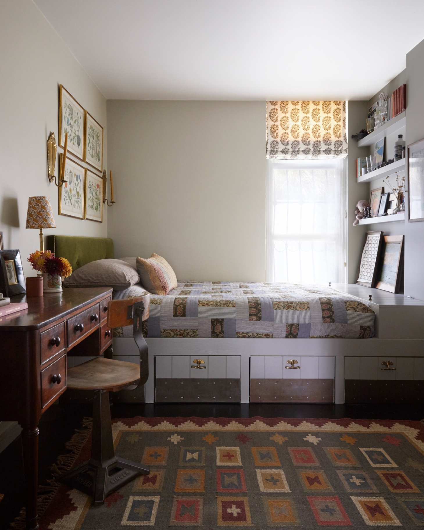 a bedroom with a built in bed with drawers underneath, a patchwork bedspread and an olive green headboard, a dark wood desk next to the bed with a stool and an earthy multicoloured rug on the floor