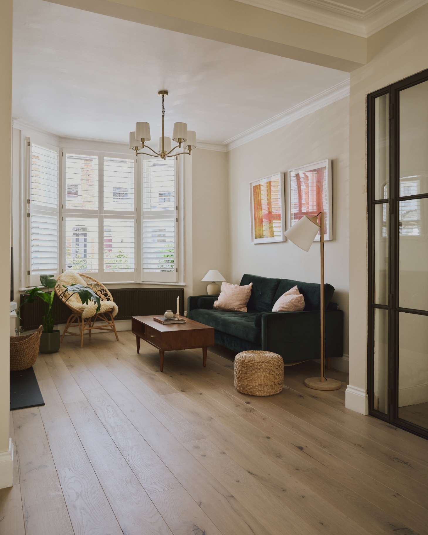a living room with hard wood floors, a dark green velvet sofa, a chandellier with gold hardware and cream shades, a large bay window with shutters and a cane armchair in front of it