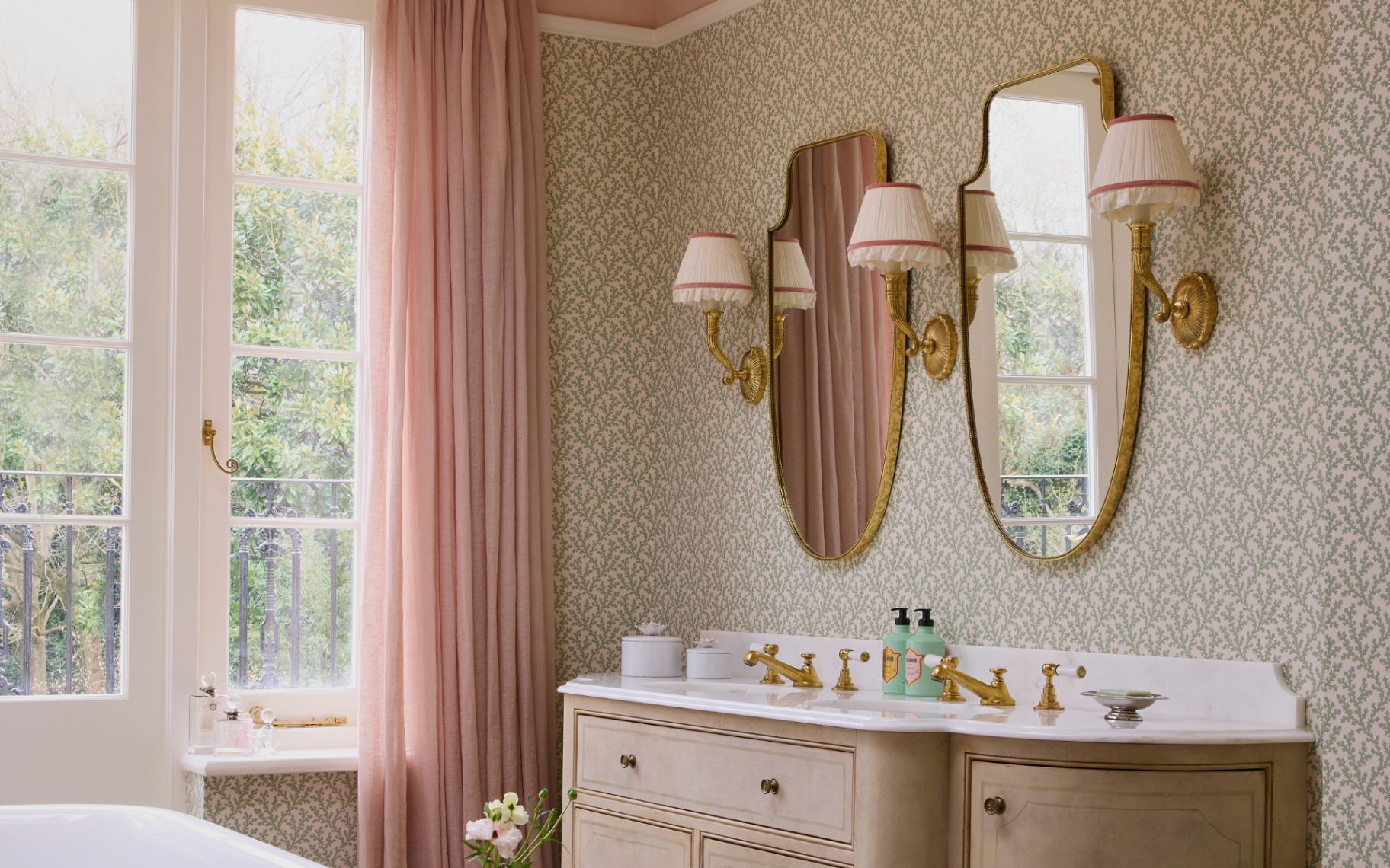 a gorgeous and warm bathroom pictured enveloped in wallpaper and dusky pink accents with an antique dresser his and hers sinks and sconces adorning the walls of a beautifully lit room