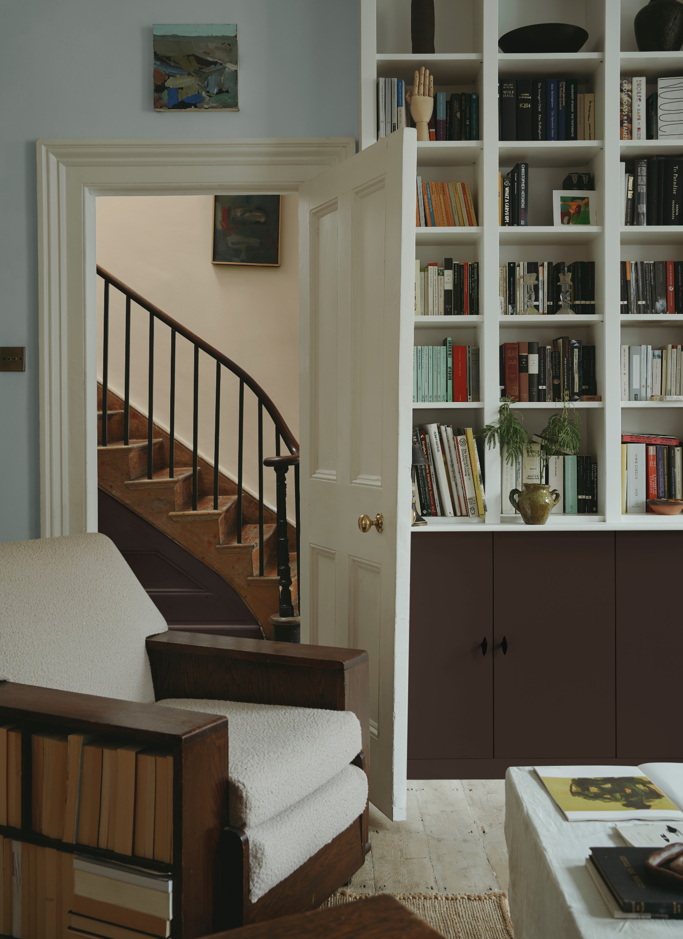 A serene room featuring white built-in shelving over deep brown cabinets, an open cream doorway revealing a wooden staircase, and an armchair with built-in book storage in its wooden armrest.
