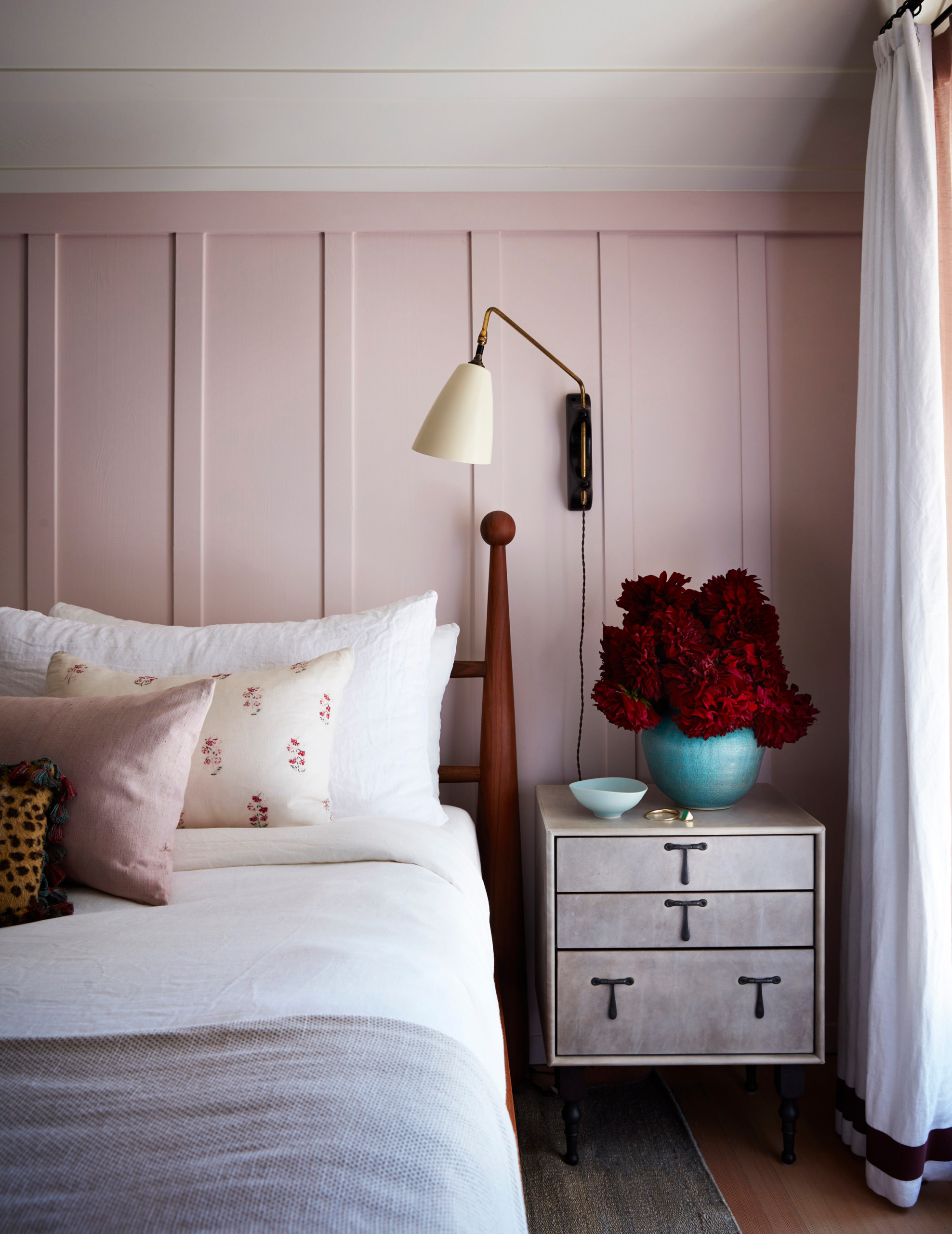 anne hathaway's bedroom, with pink panelled walls, a large wall light, a bed with white bedding and a floral cushion and a grey bedside table with a turqoise vase with a bunch of red flowers