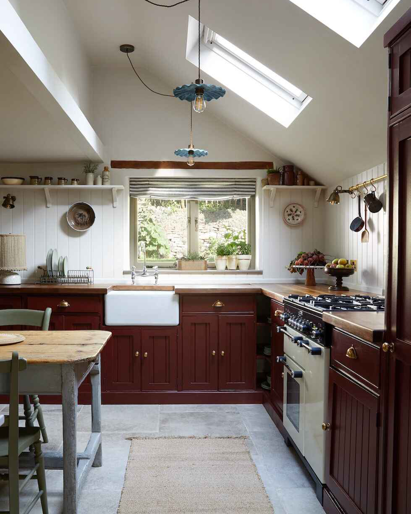 a kitchen with dark red painted cabinets, blue frilled pendants, a cream range cooker and a grey rustic dining table