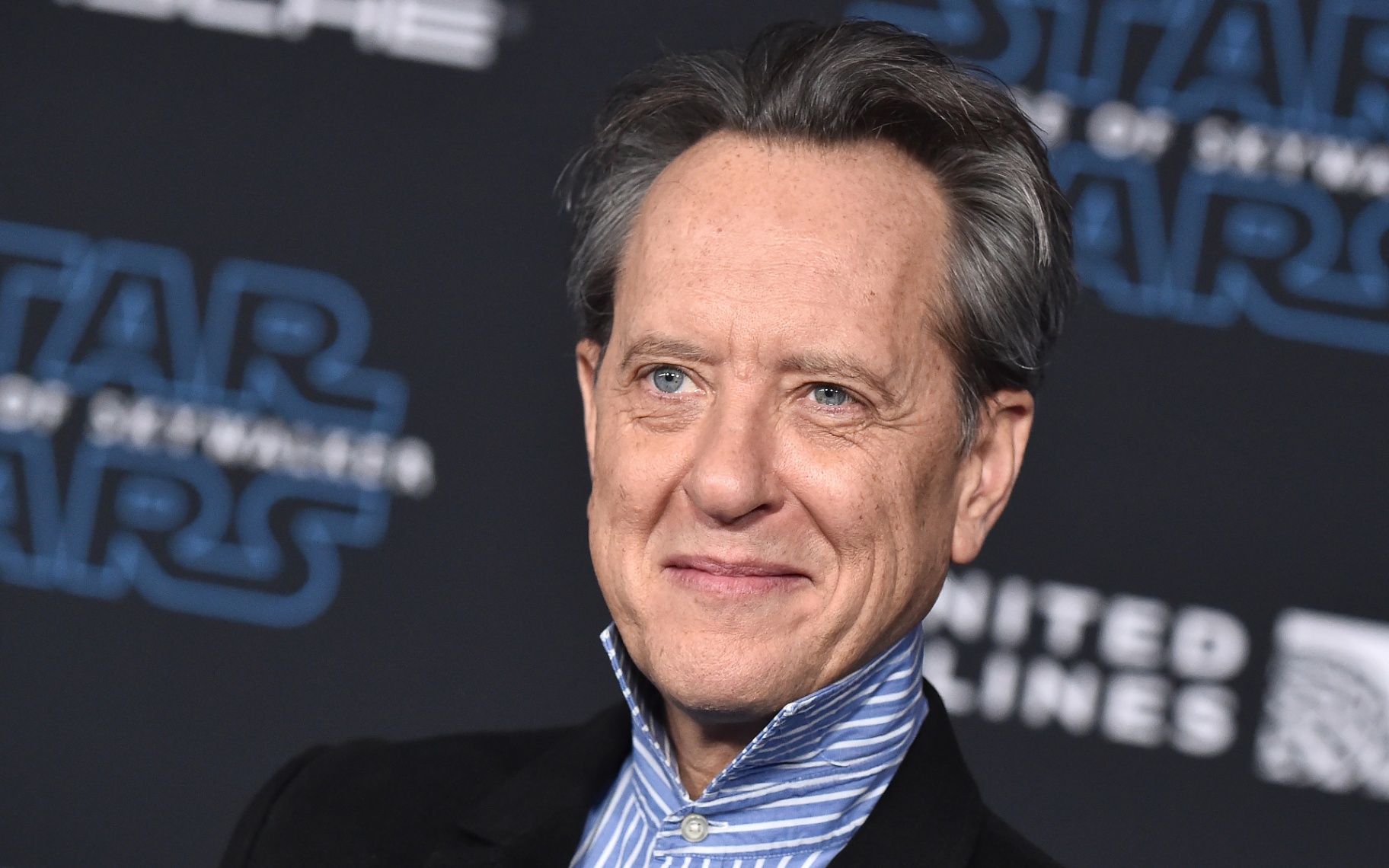 Actor Richard E. Grant smiling at a film premiere, wearing a black blazer with a blue and white striped popped-collar shirt against a dark Star Wars backdrop