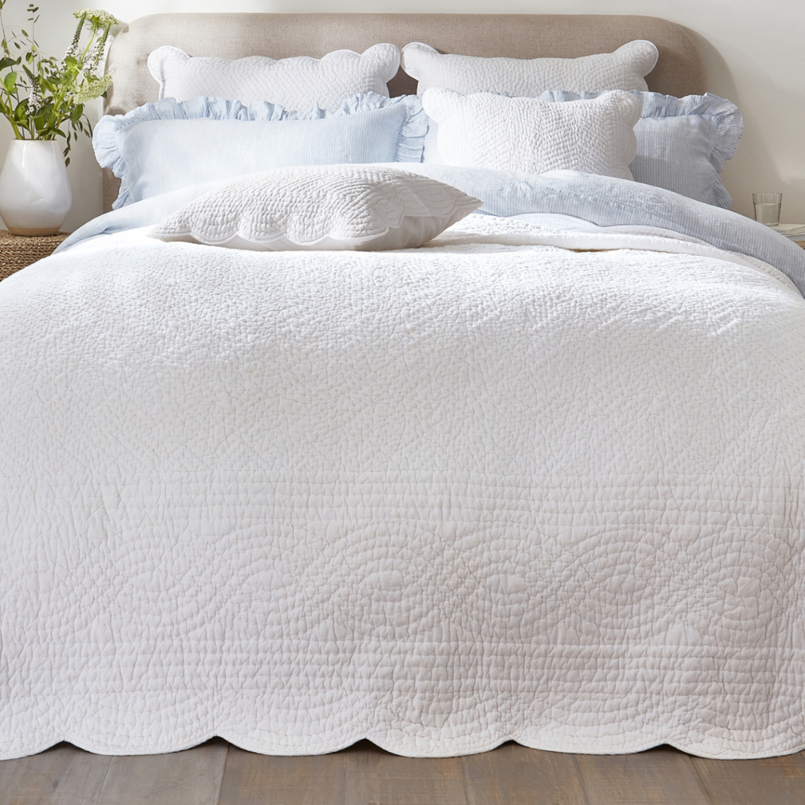 a white stitch patterned bedspread with a scalloped trim on a bed with blue ruffled cushions