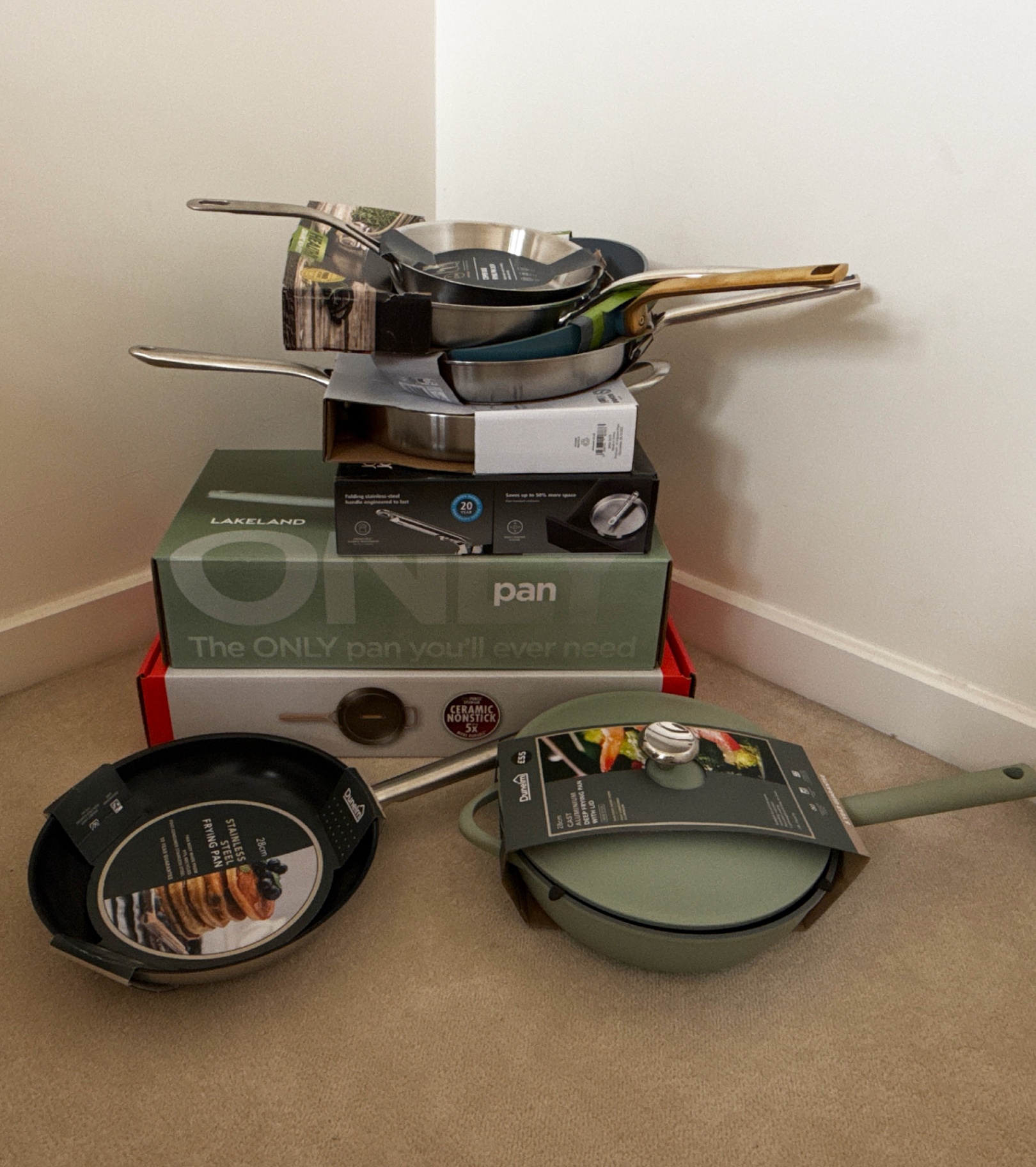 a pile of 11 frying pans in their packaging on a cream carpet against a white wall